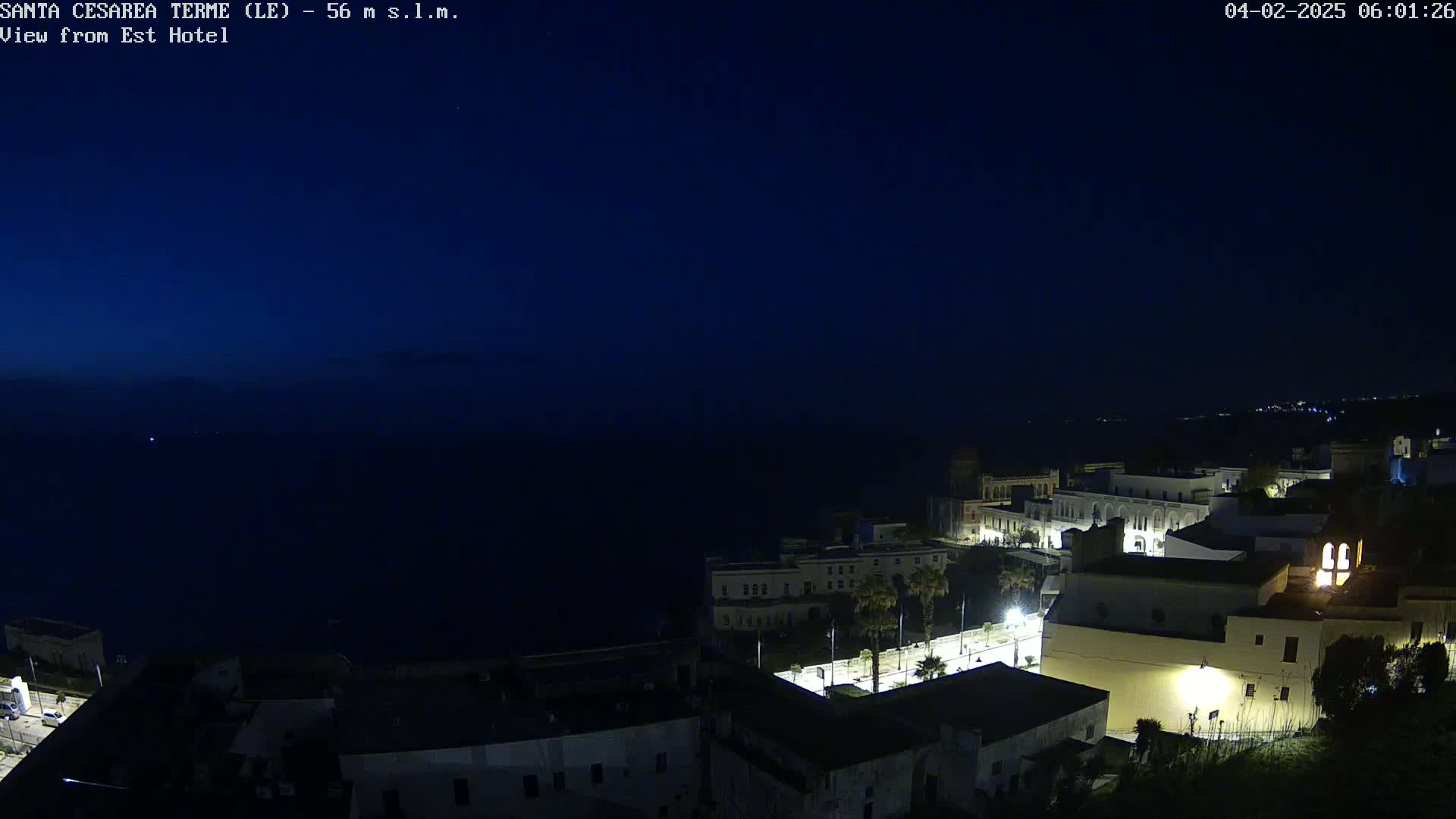 A nighttime, coastal town with illuminated buildings overlooks a dark sea under a clear, dark blue sky.