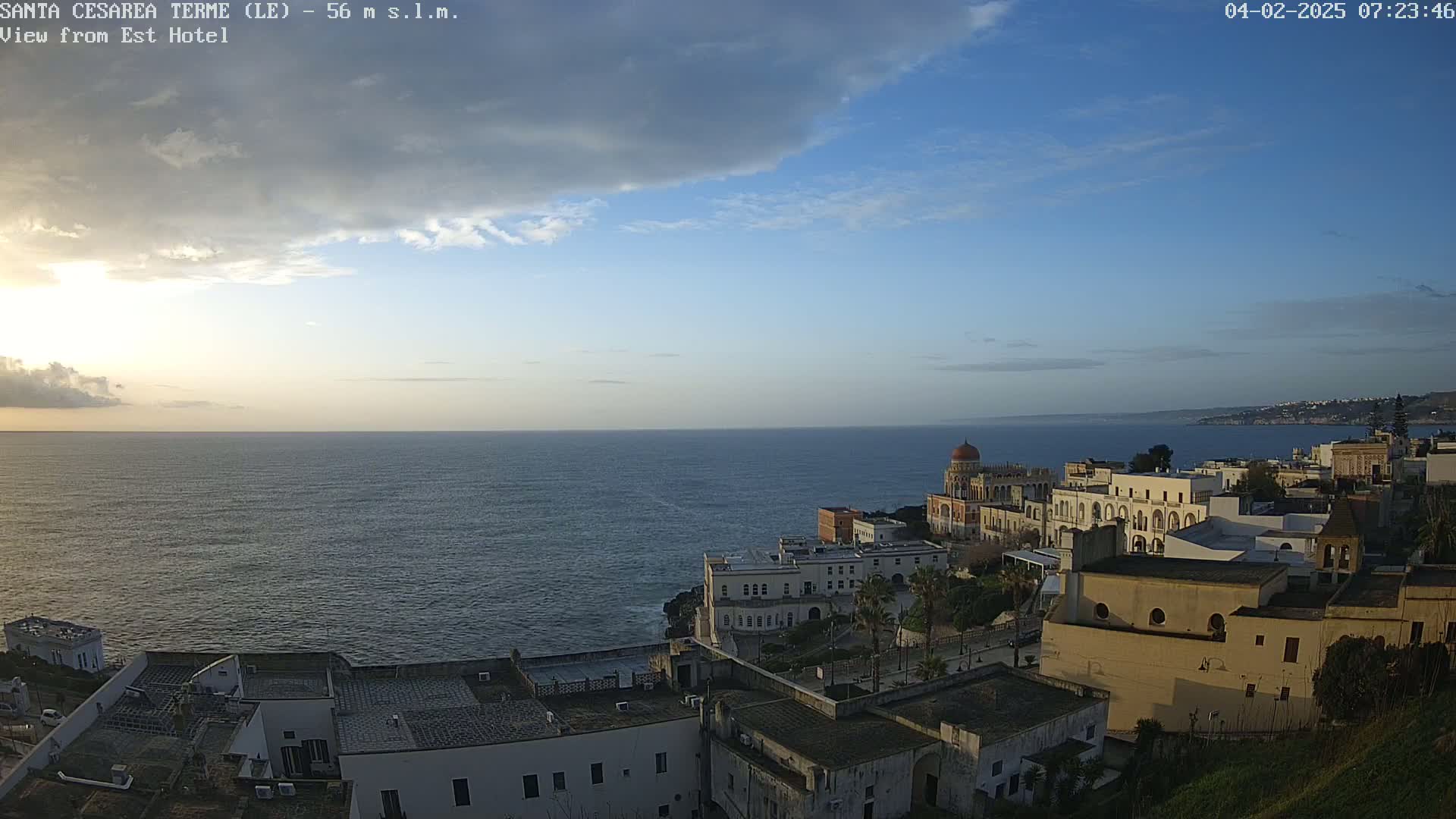 A coastal town with white buildings overlooks a calm sea under a partly cloudy sky.