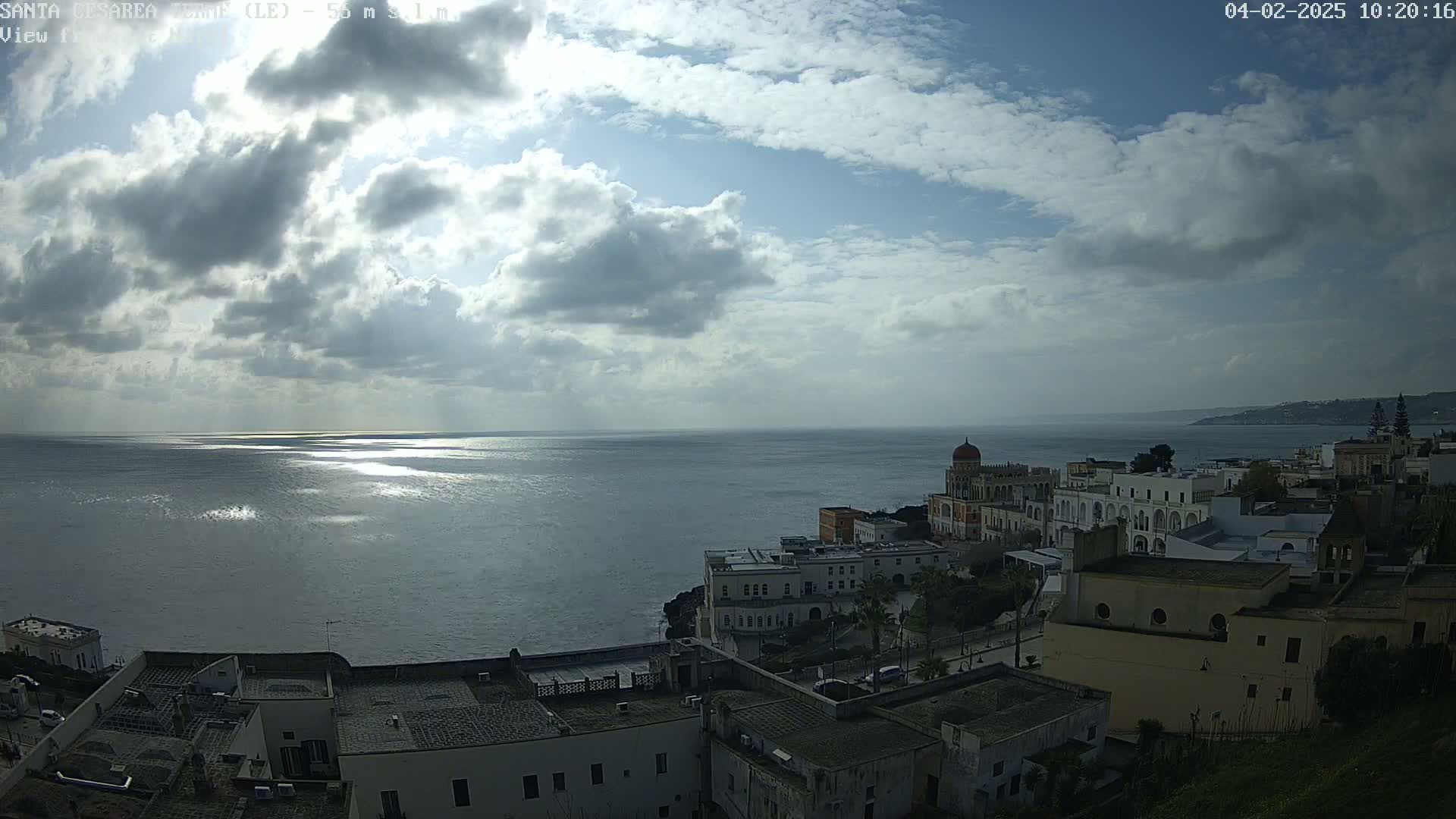 A partly cloudy day reveals a coastal town built on a cliff overlooking a calm sea, with sunlight reflecting on the water's surface.