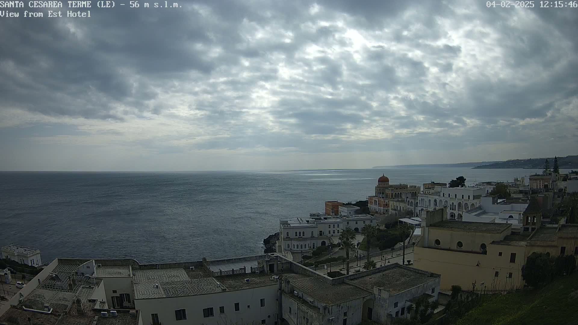 A coastal town with white buildings and a domed structure overlooks a calm, dark blue sea under a mostly cloudy sky.