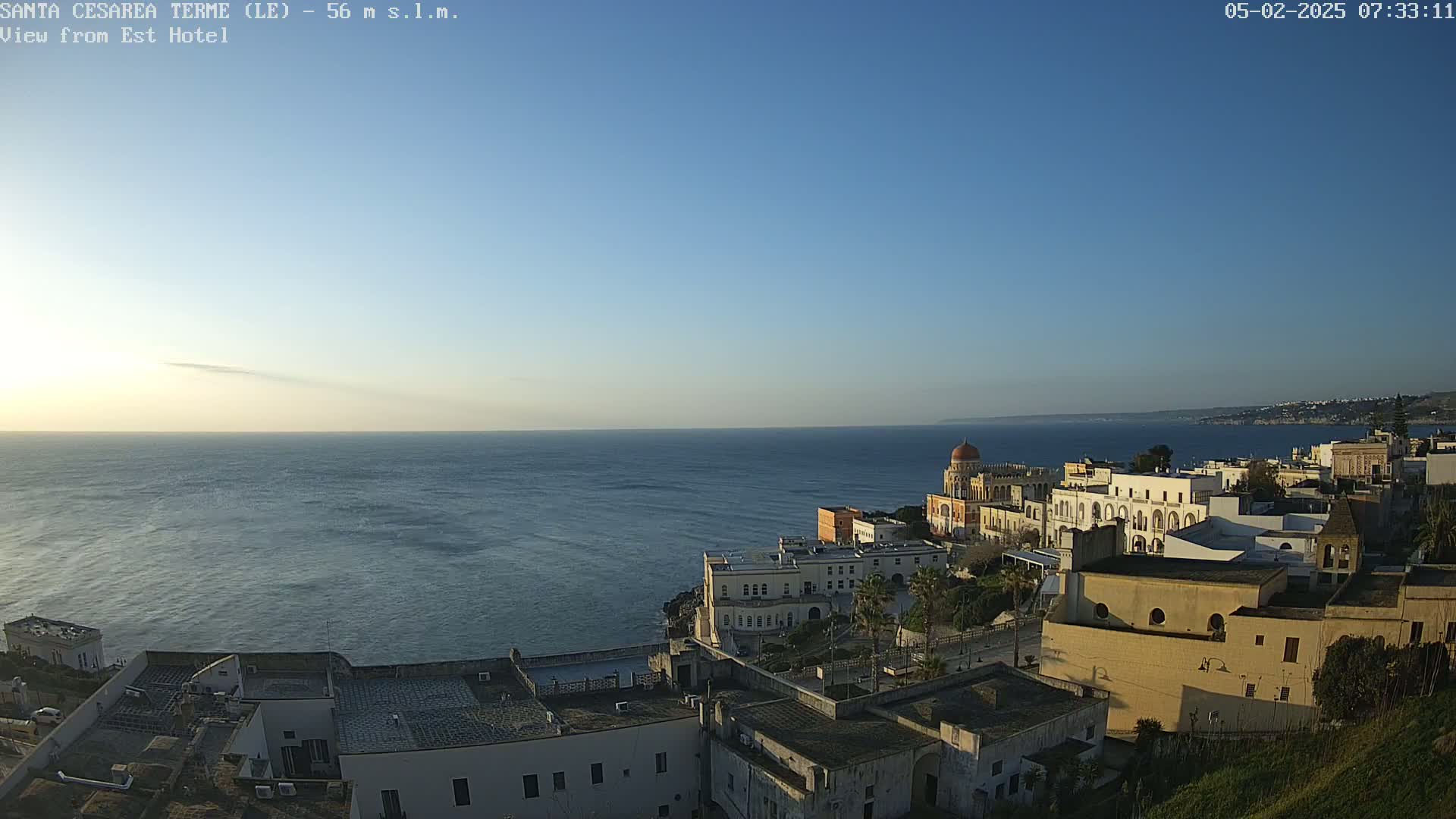 A coastal town with white buildings and a domed structure overlooks a calm, blue sea under a clear blue sky.