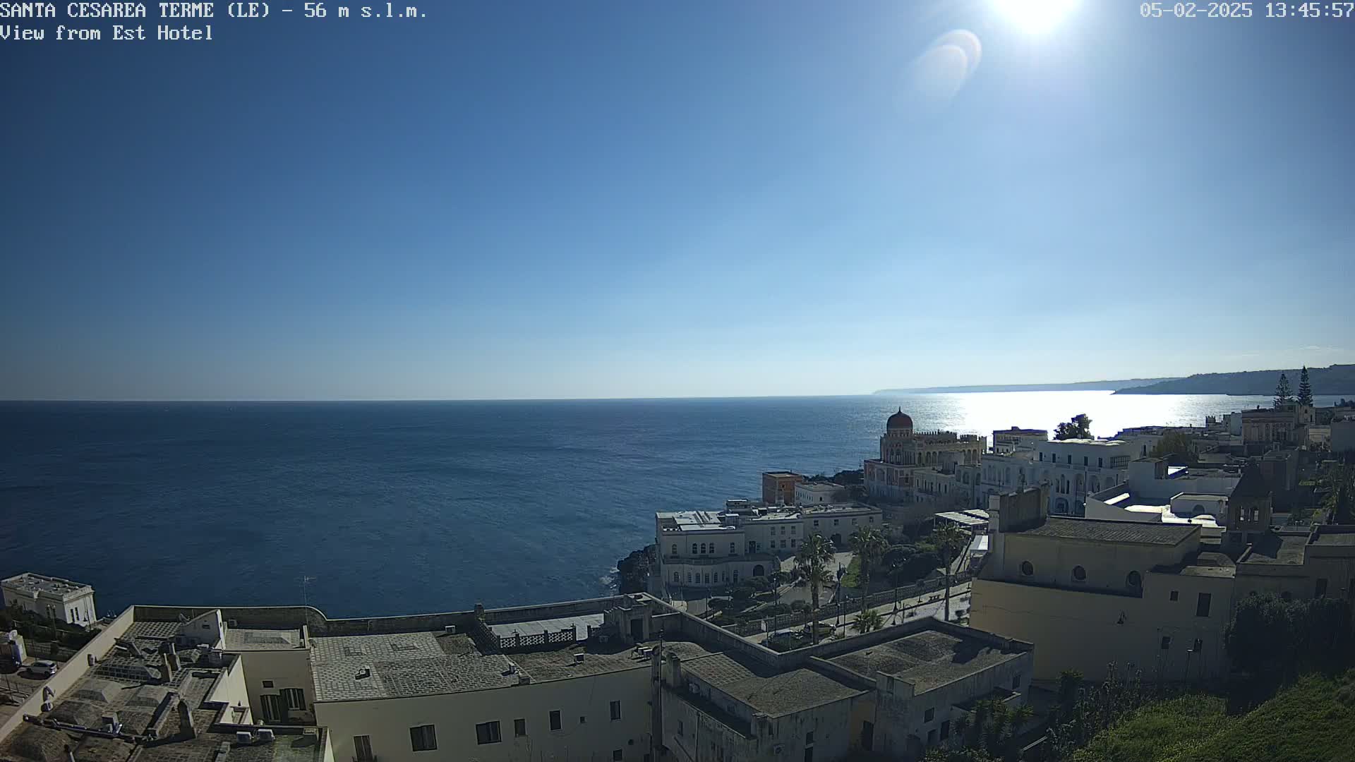 A coastal town with white buildings sits on a cliff overlooking a calm, blue sea under a clear, sunny sky.