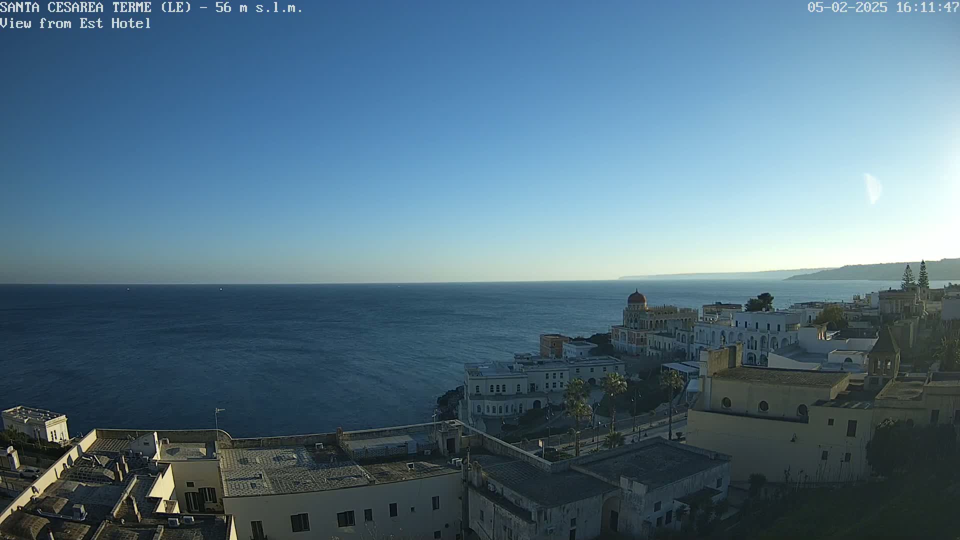 A coastal town with white buildings overlooks a calm, dark blue sea under a clear, bright blue sky.