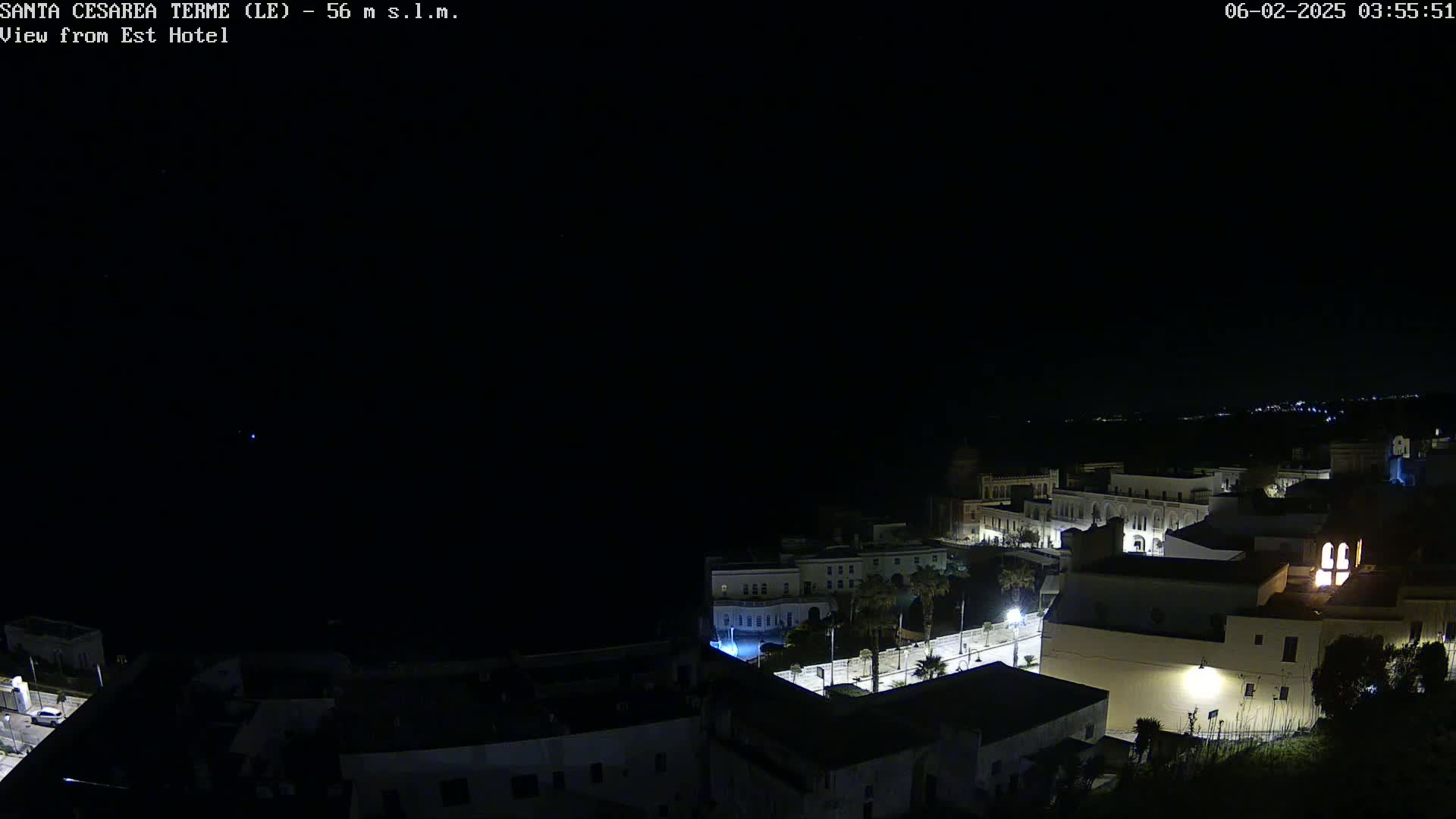 A nighttime, high-angle view of a coastal town with dimly lit buildings, streets, and palm trees under a clear, dark sky.