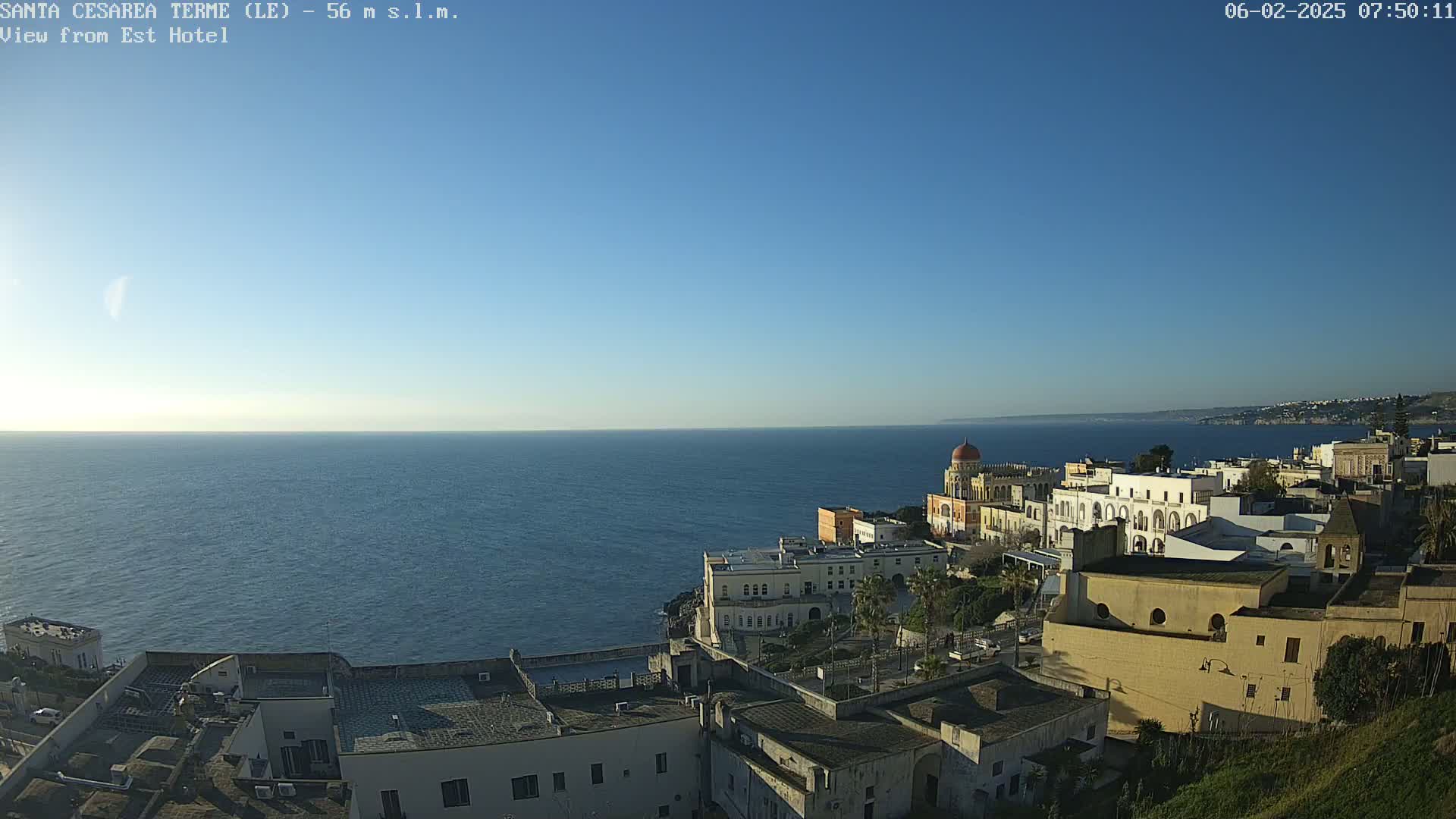A clear, sunny day overlooks a coastal town with buildings cascading down a hillside toward a calm sea.