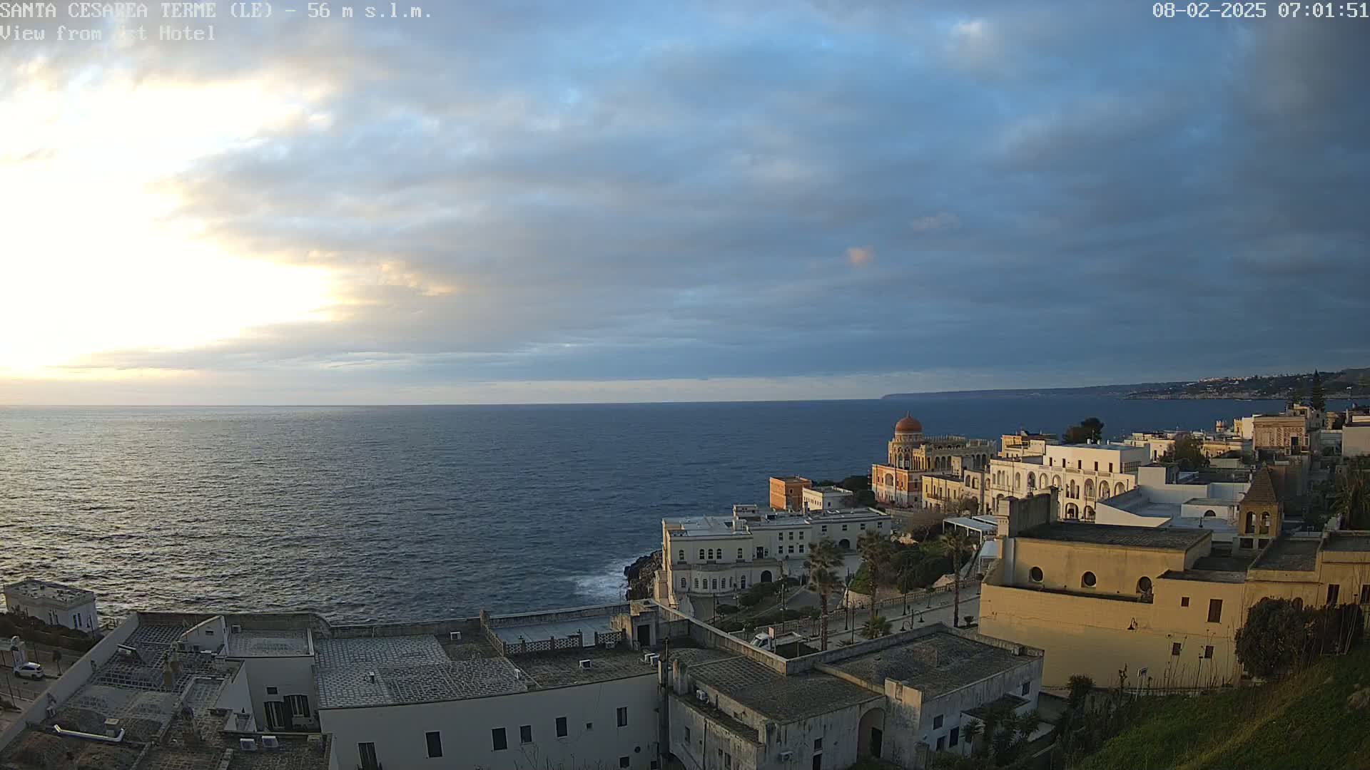 A coastal town with white buildings, some with domes, sits on a cliff overlooking a calm, dark blue sea under a mostly cloudy sky with a hint of sunrise.