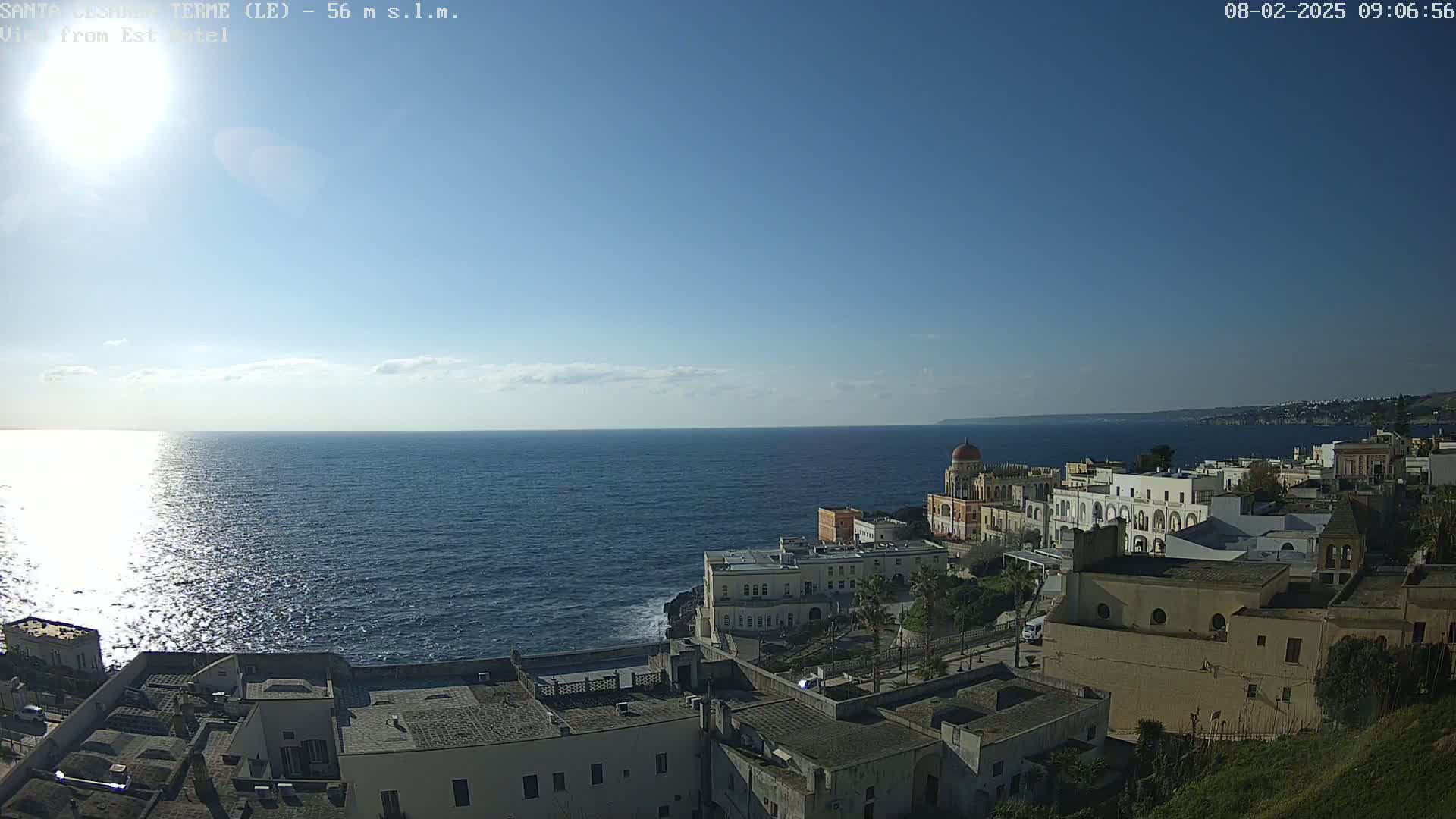 A sunny day reveals a coastal town nestled against a calm, blue sea under a clear sky.