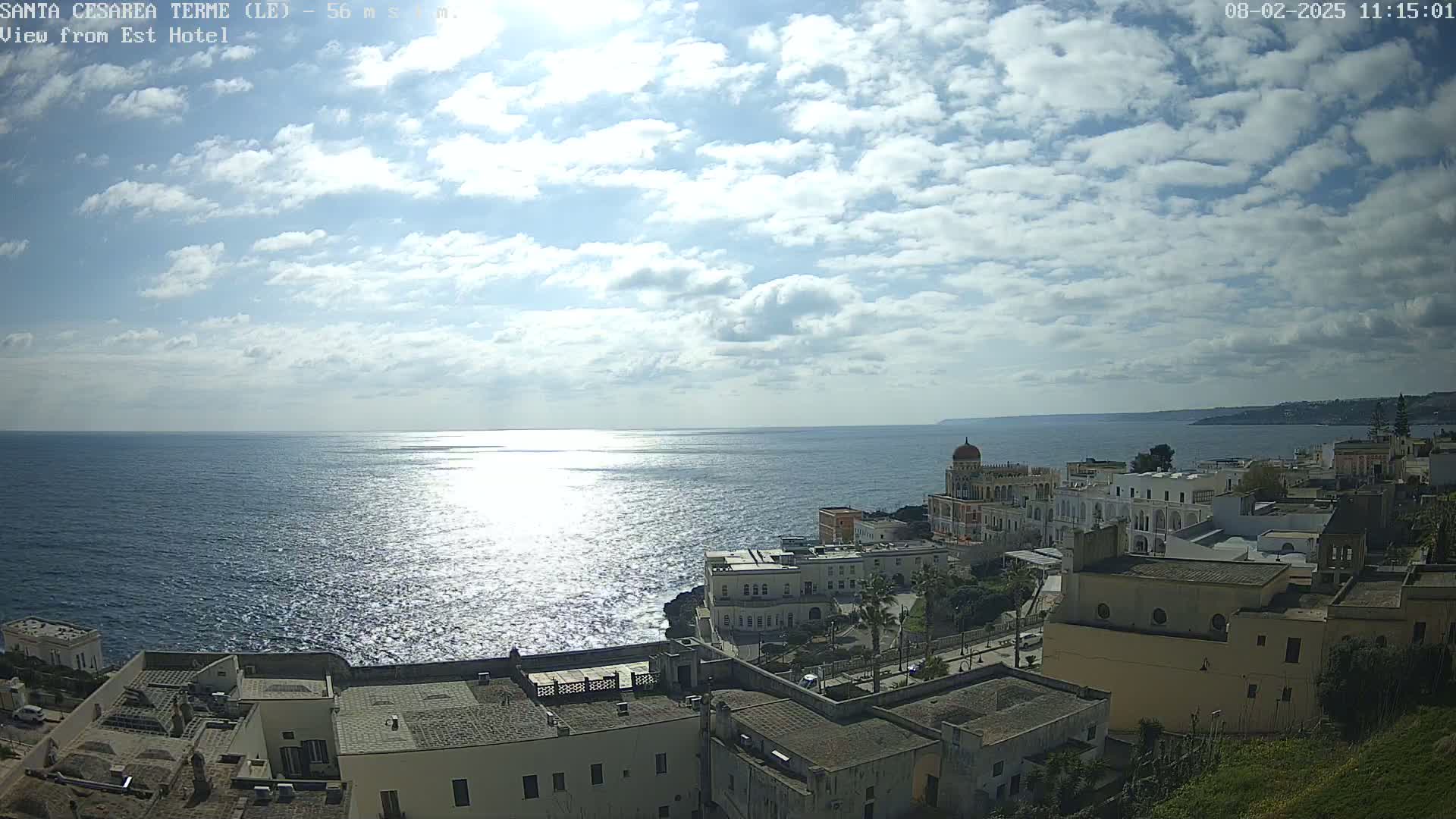 A coastal town with white buildings sits on a cliff overlooking a sparkling sea under a partly sunny sky with scattered clouds.