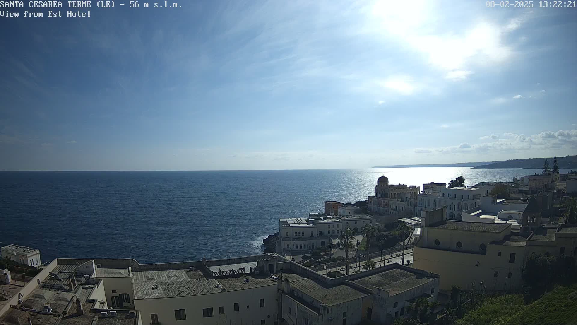 A coastal town with white buildings sits on a rocky outcrop overlooking a calm, sunny sea under a mostly clear sky.