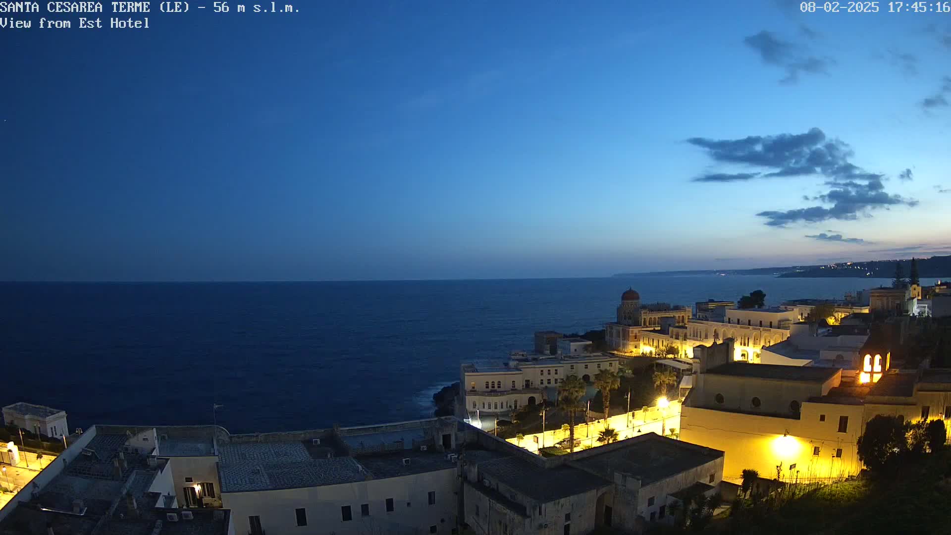 A coastal town at twilight is illuminated, overlooking a dark blue sea under a mostly clear sky with a few clouds.