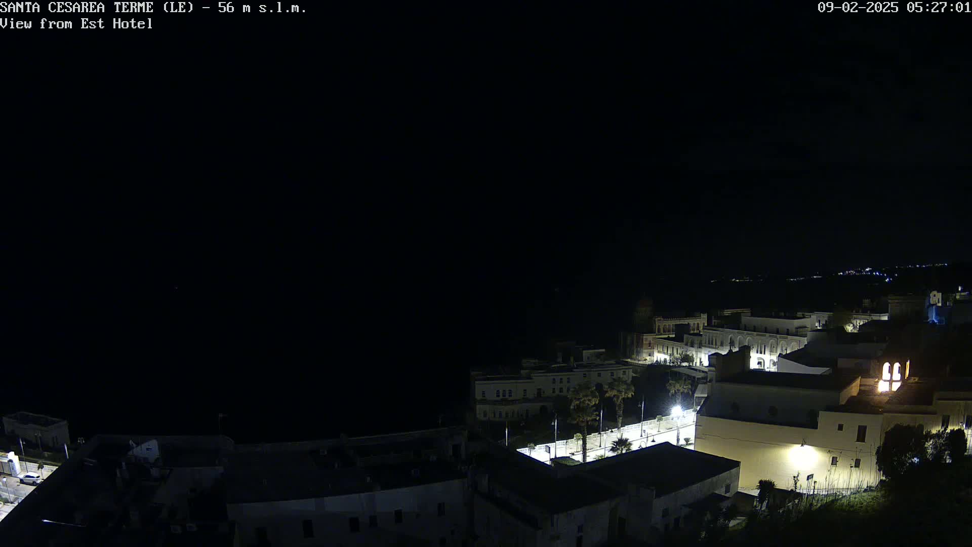 A nighttime, high-angle view of a coastal town with dimly lit buildings and streets, overlooking a dark ocean under a clear, starless sky.