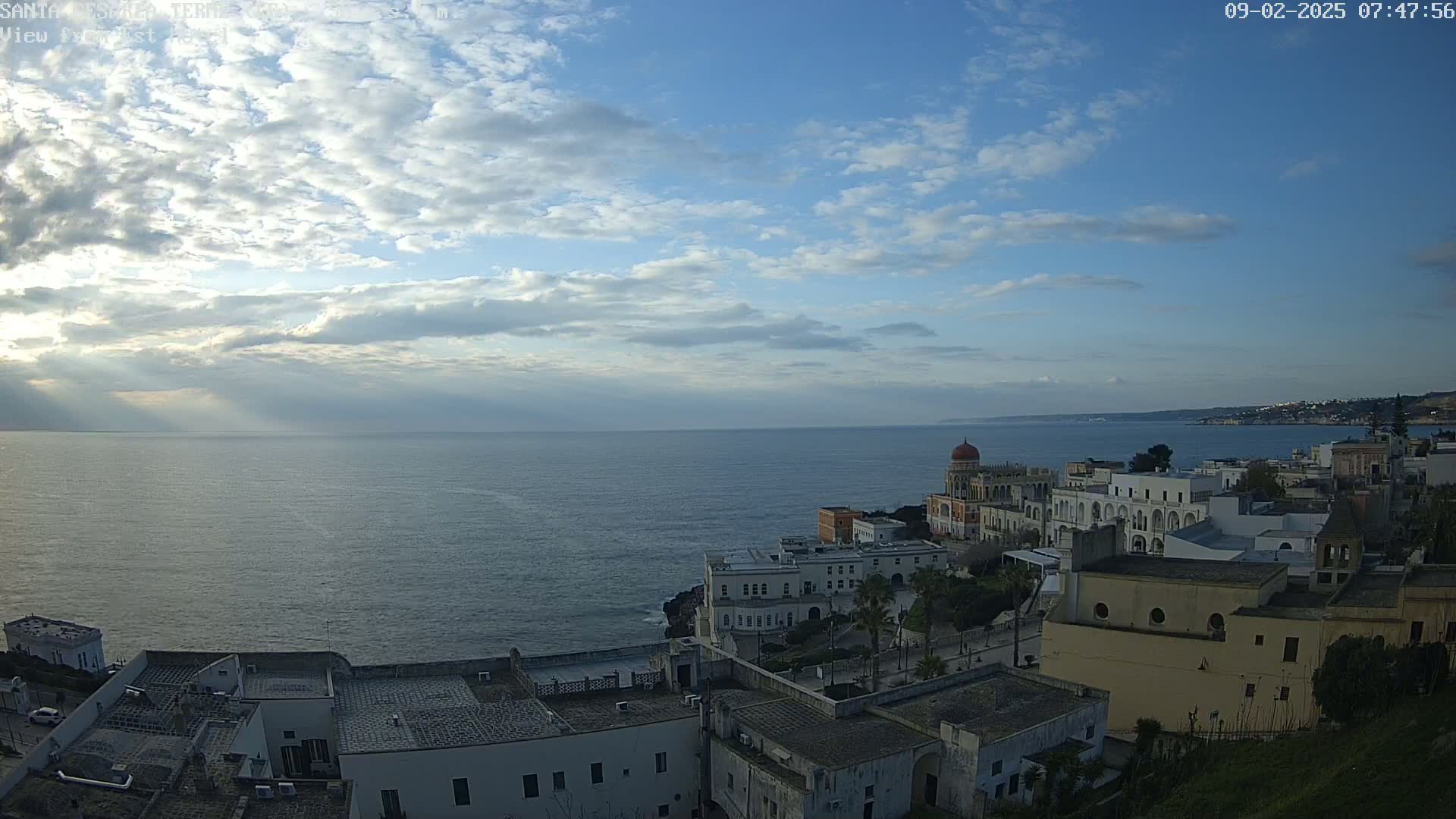 A coastal town with white buildings and a red-domed structure overlooks a calm sea under a partly cloudy sky with sun rays breaking through the clouds.