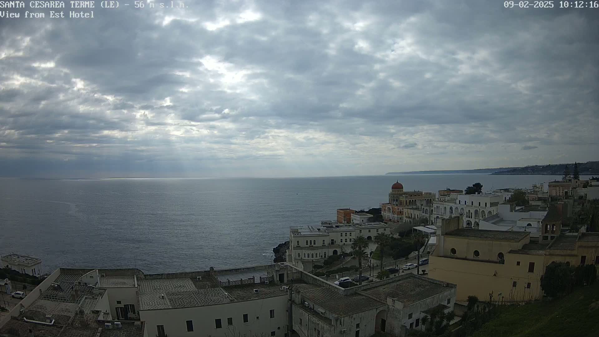 A coastal town with white buildings sits on a cliff overlooking a calm sea under a cloudy sky.