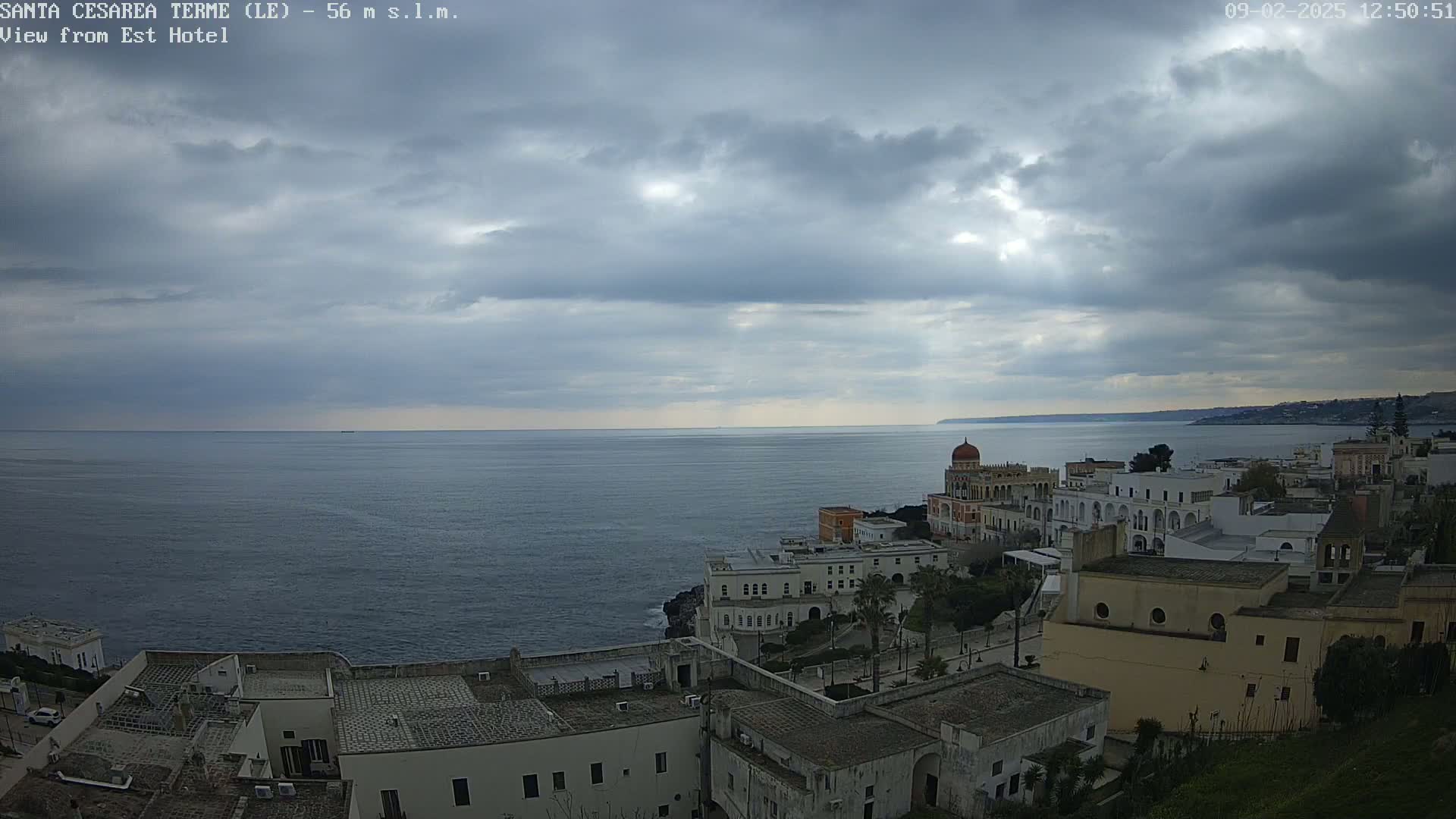 A coastal town with white buildings sits on a cliff overlooking a calm, dark-blue sea under a mostly cloudy sky.