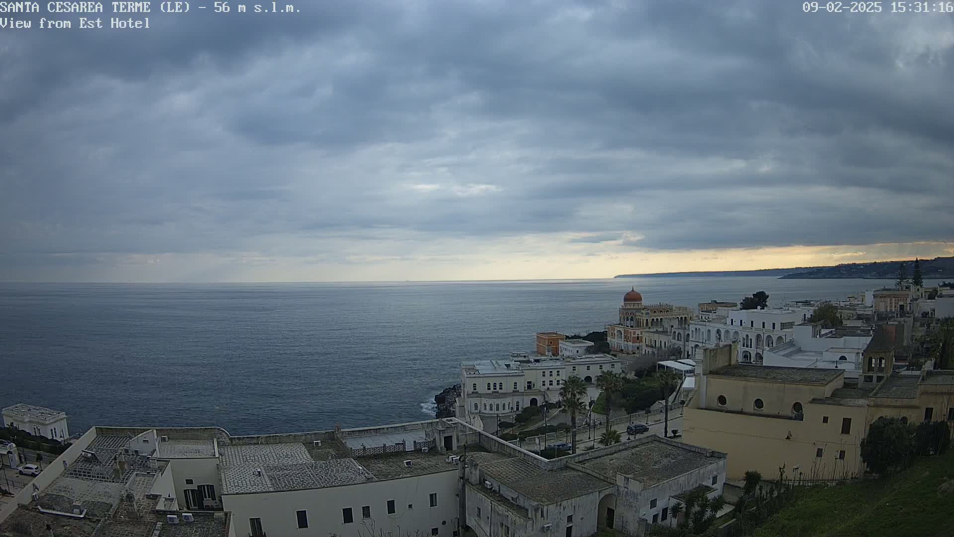 A coastal town with white buildings overlooks a calm, dark blue sea under a mostly cloudy sky.