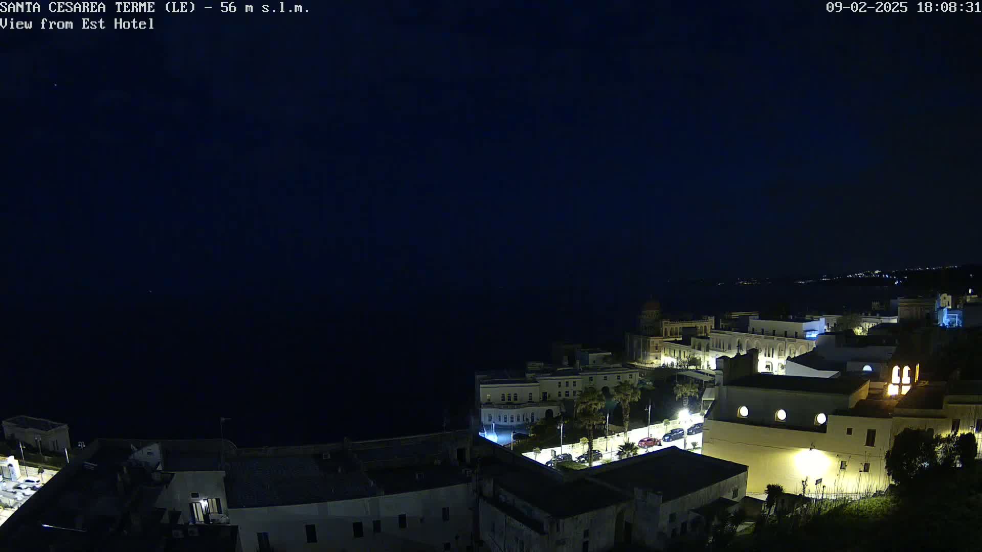 A nighttime view of a coastal town with buildings illuminated, overlooking a dark sea under a clear, dark sky.