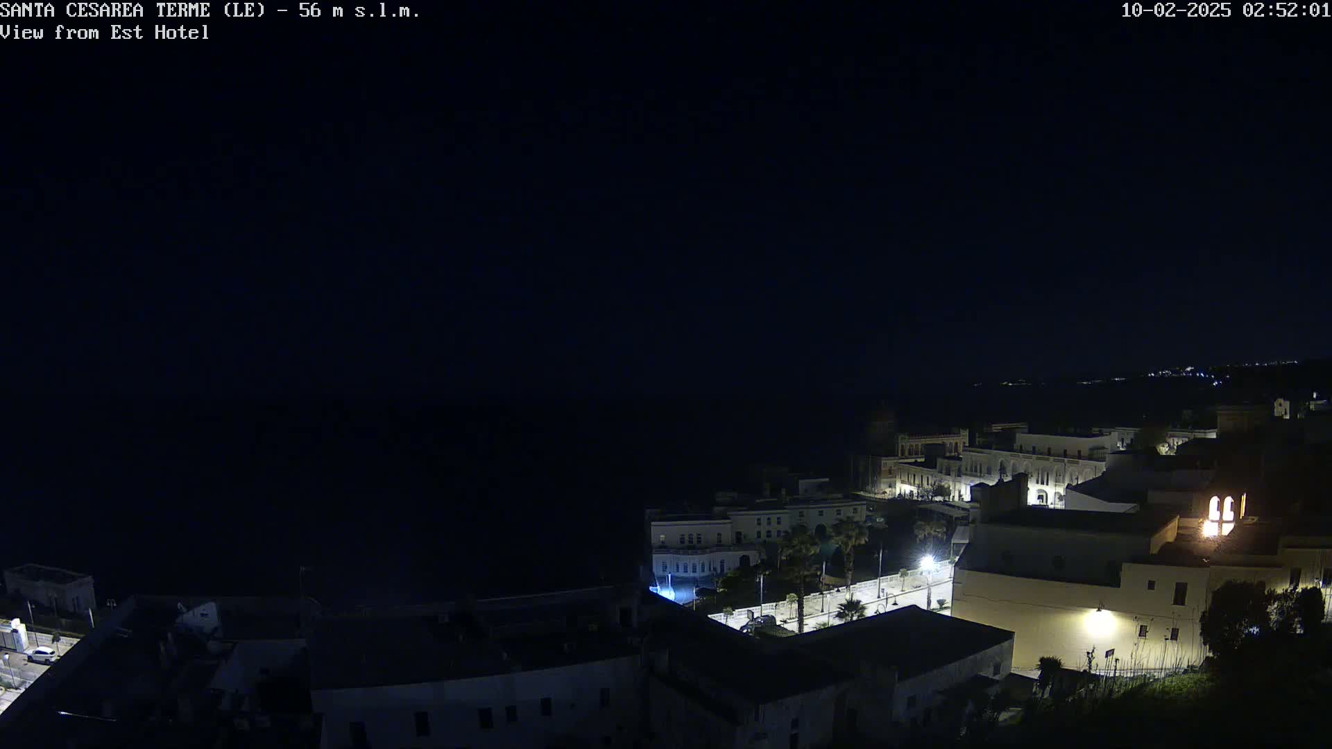 A nighttime, aerial view of a coastal town with dimly lit buildings, overlooking a dark body of water under a clear, dark sky.