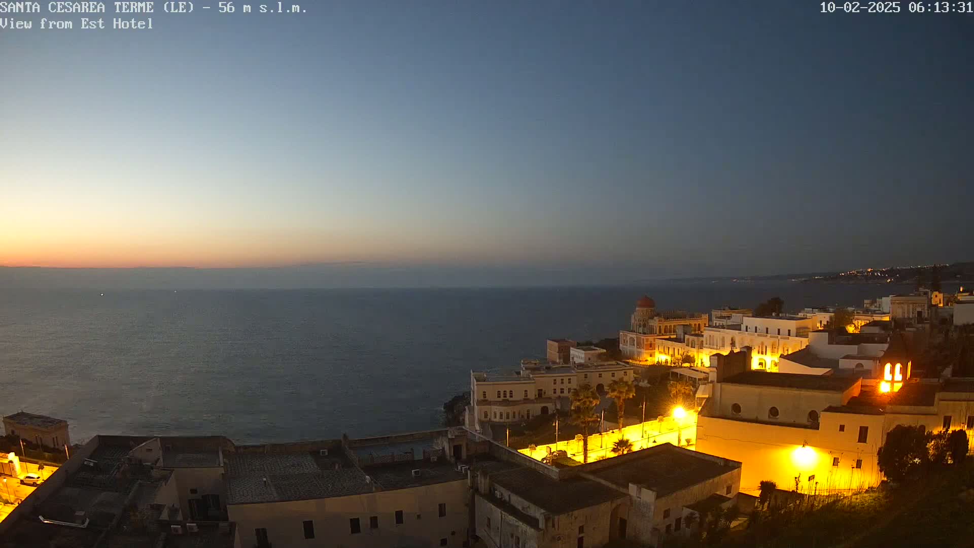 A coastal town is illuminated at twilight, overlooking a calm sea under a clear, dark blue sky.