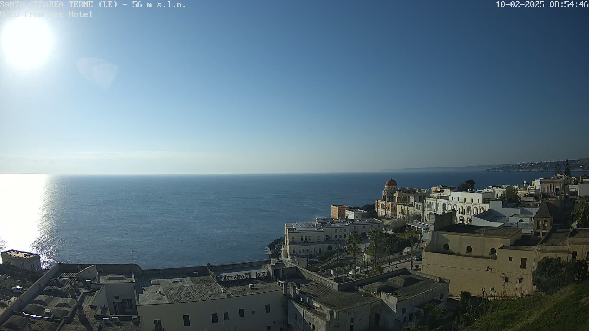 A coastal town with white buildings sits on a cliff overlooking a calm, sunny ocean under a clear blue sky.