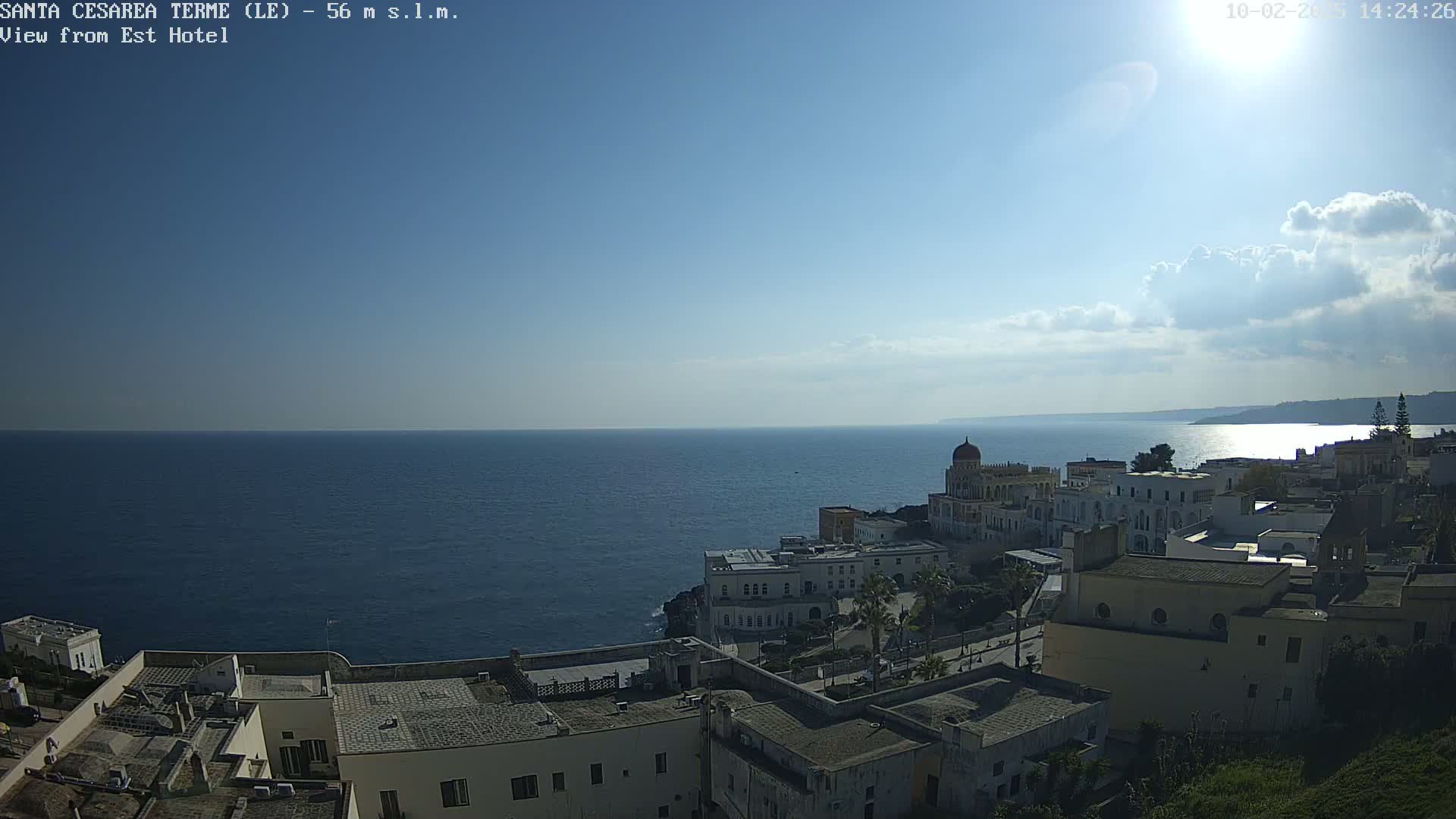 A coastal town with white buildings overlooks a calm blue sea under a mostly sunny sky with some scattered clouds.