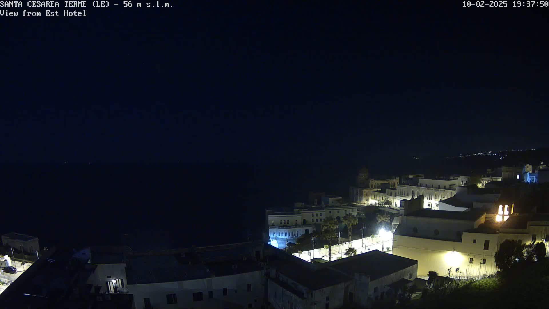 A nighttime, high-angle view of a coastal town with dimly lit buildings and a dark sea in the background under a clear, dark sky.