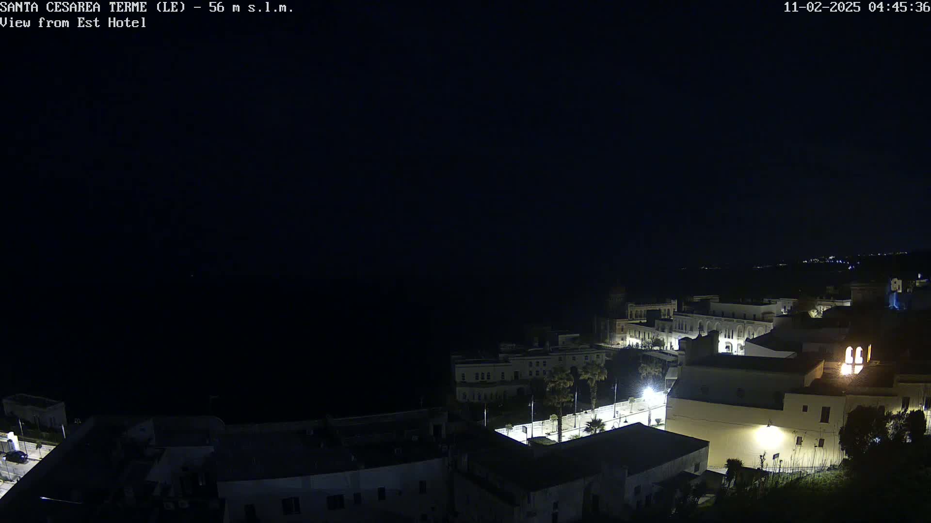 A nighttime, long shot of a coastal town with dimly lit buildings, overlooking a dark body of water under a clear, black sky.