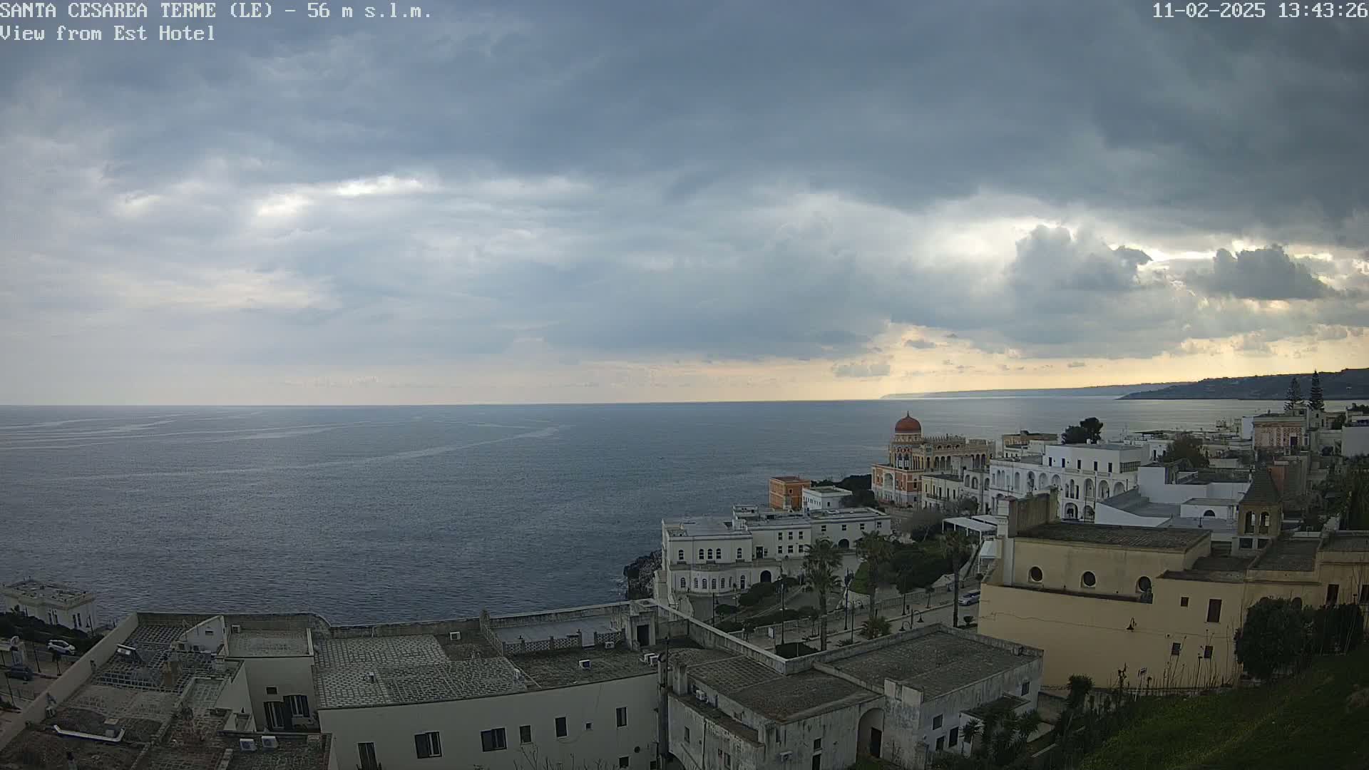 A coastal town with white buildings sits on a cliff overlooking a calm sea under a mostly cloudy sky.