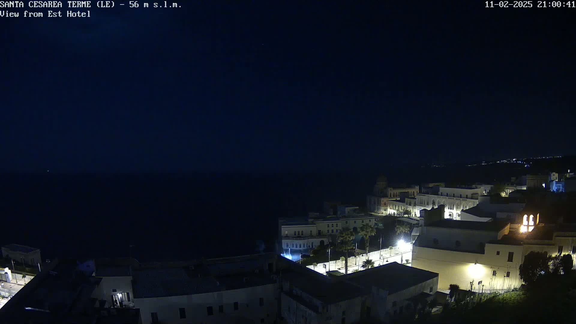A nighttime coastal town with illuminated buildings overlooks a dark ocean under a clear, dark sky.