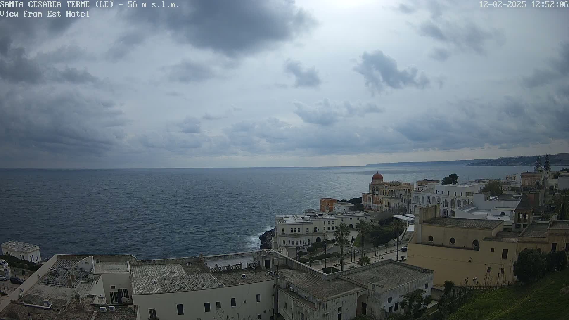 A coastal town with white buildings and a domed structure sits on a cliff overlooking a dark blue sea under a mostly cloudy sky.