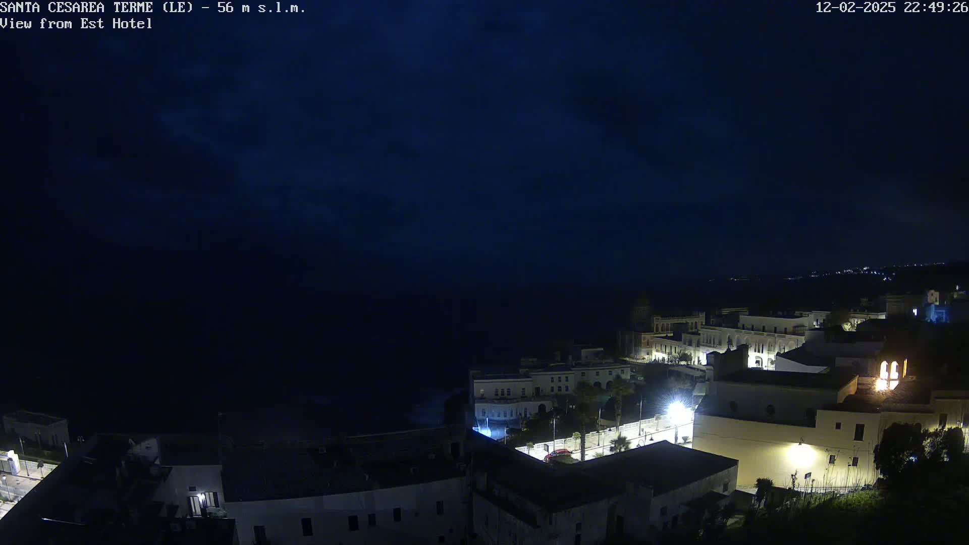 A nighttime coastal town is dimly lit under a dark, cloudy sky, with the sea visible in the background.