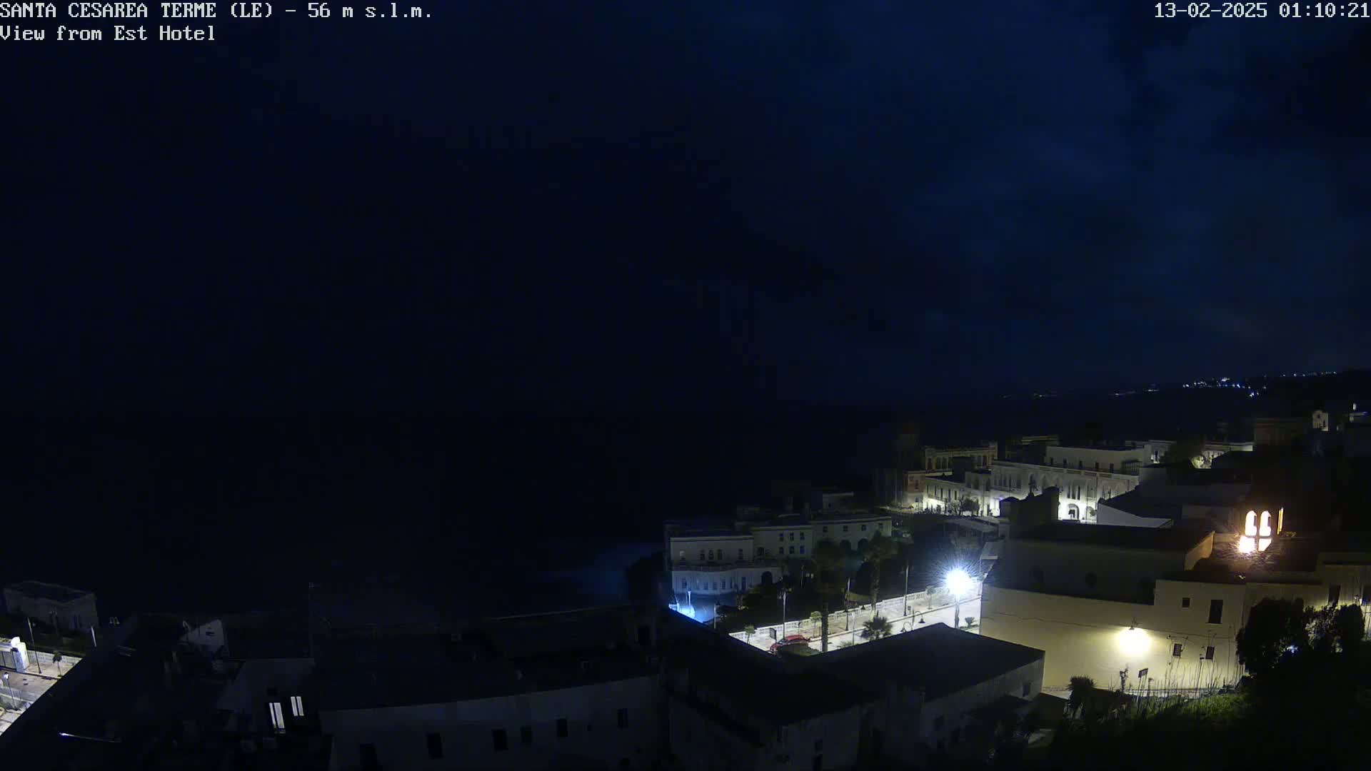 A nighttime coastal town is dimly lit, with a dark ocean visible in the background under a mostly cloudy sky.
