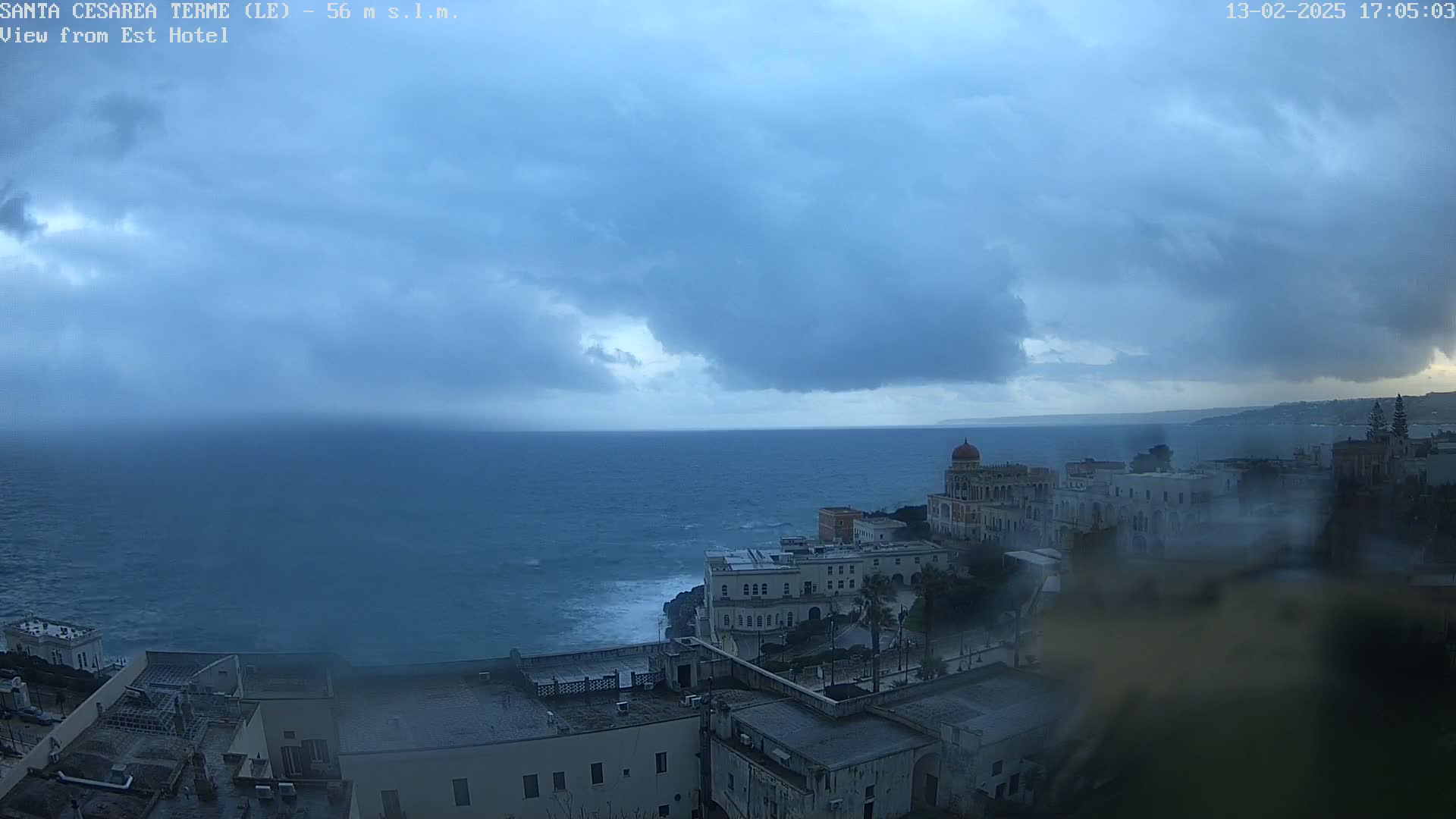 A coastal town with buildings sits under a stormy, overcast sky, overlooking a choppy sea.