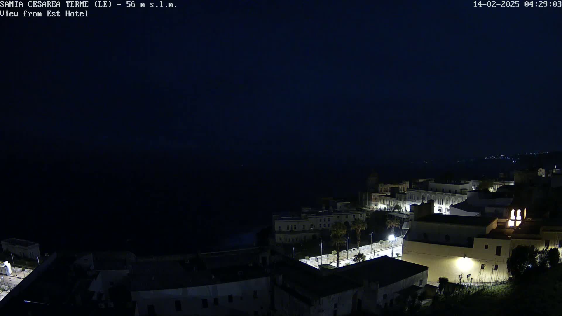 A dark, nighttime coastal town is illuminated by scattered lights, overlooking a black sea under a clear, dark sky.