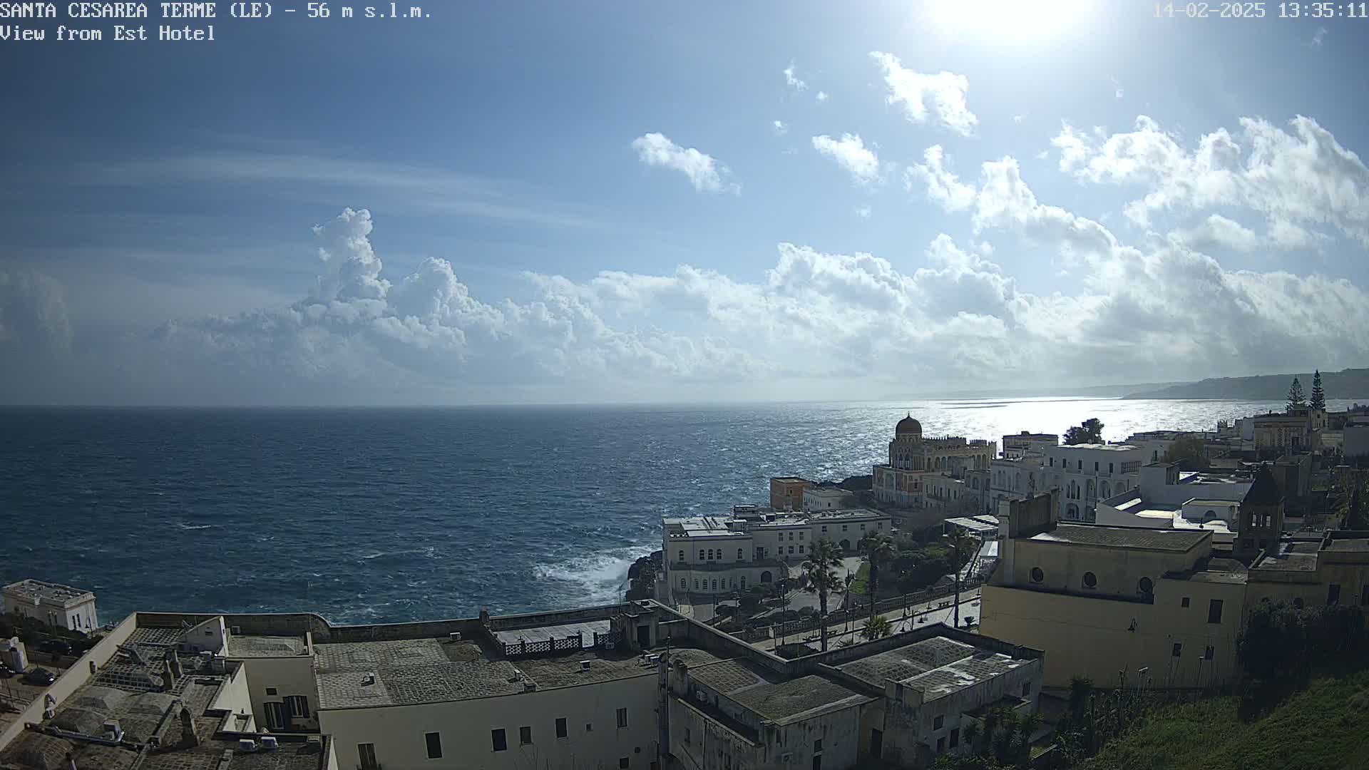 A coastal town with white buildings sits on a cliff overlooking a wavy ocean under a partly cloudy sky.