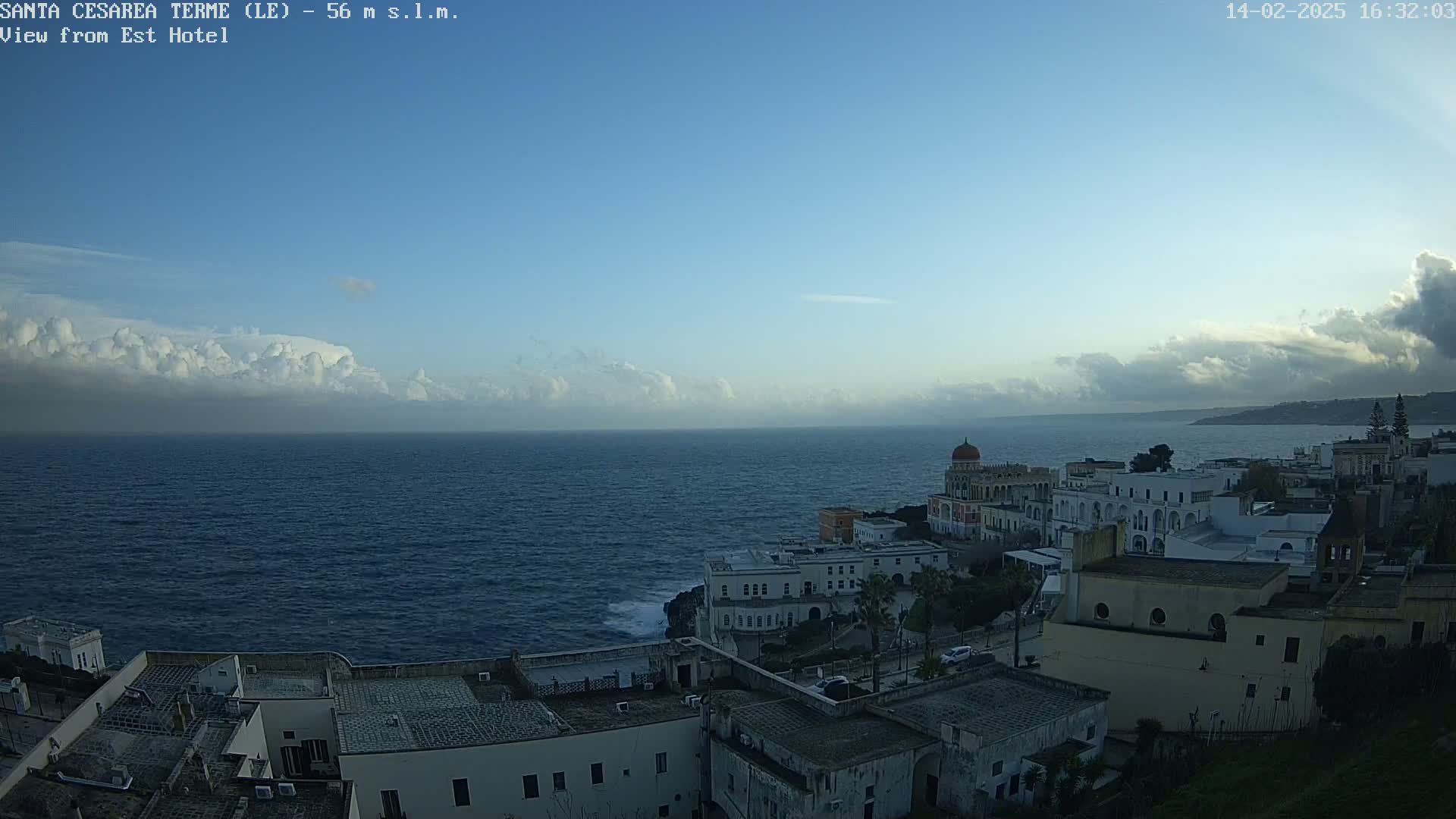 A coastal town with white buildings is situated on a cliff overlooking a dark blue sea under a mostly clear sky with a band of clouds on the horizon.