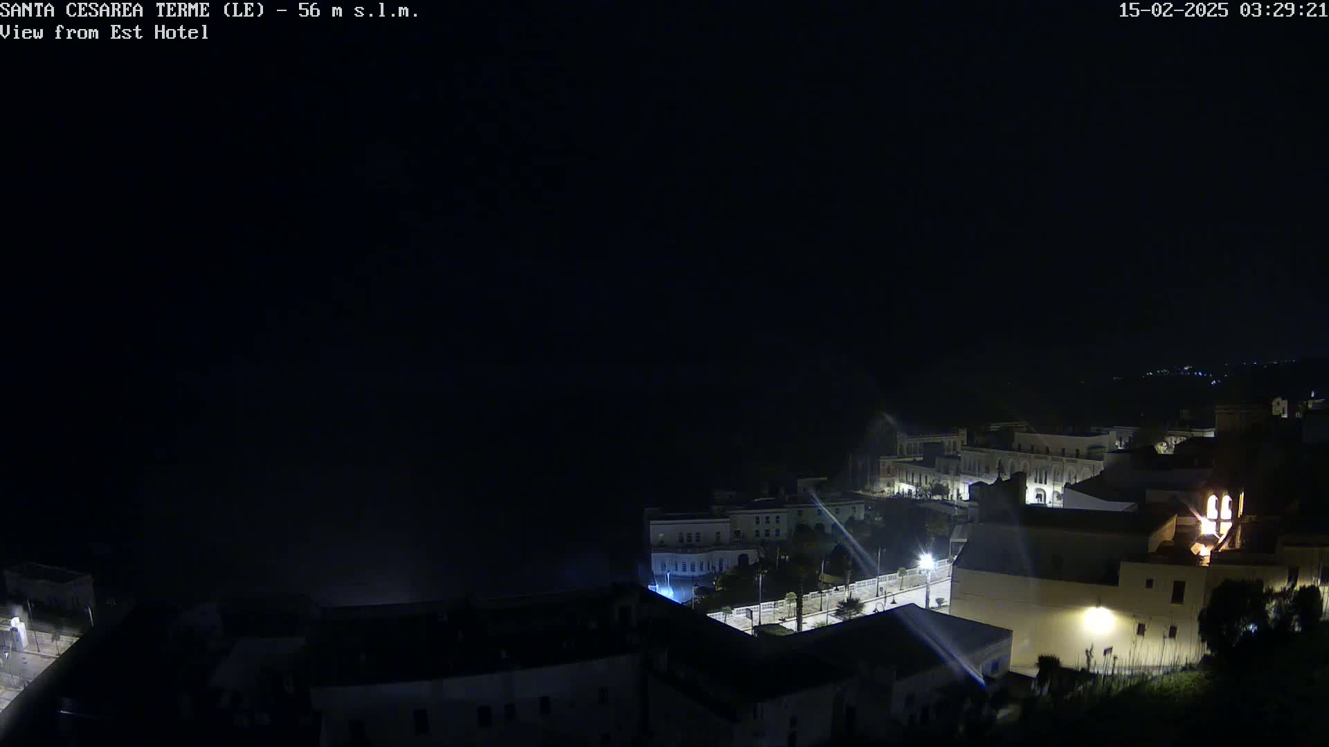 A nighttime, high-angle view of a coastal town with dimly lit buildings and a dark ocean in the background.