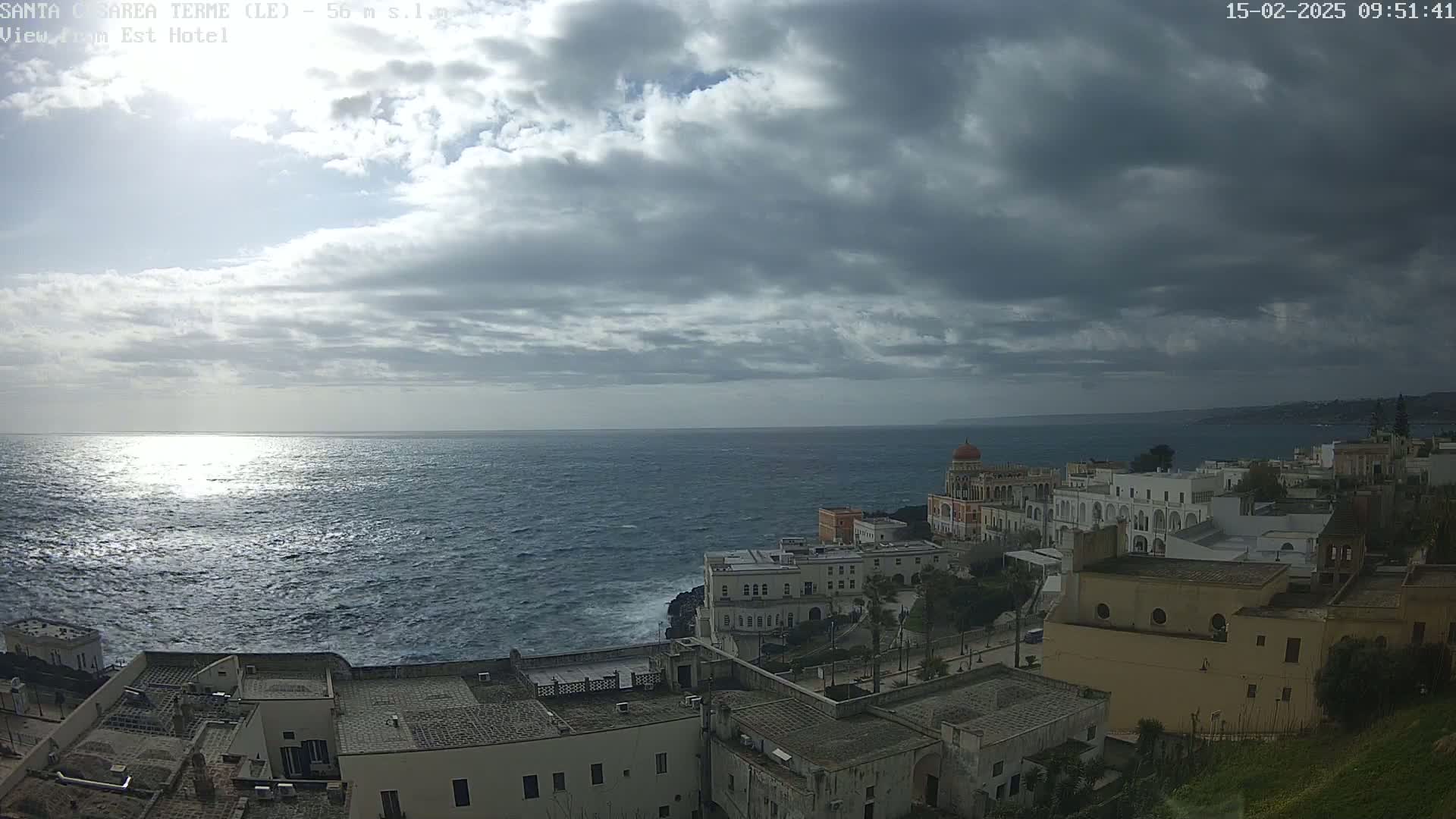 A coastal town with white buildings sits on a cliff overlooking a dark, choppy sea under a cloudy sky.