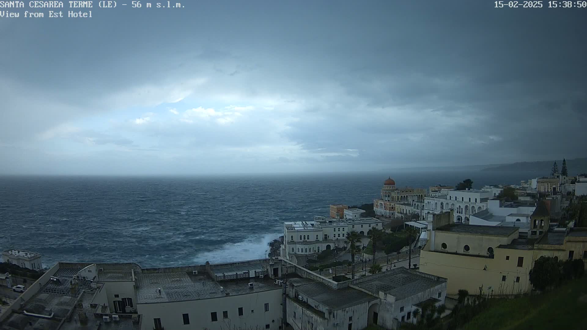 A coastal town with white buildings sits on a cliff overlooking a turbulent sea under a cloudy sky.