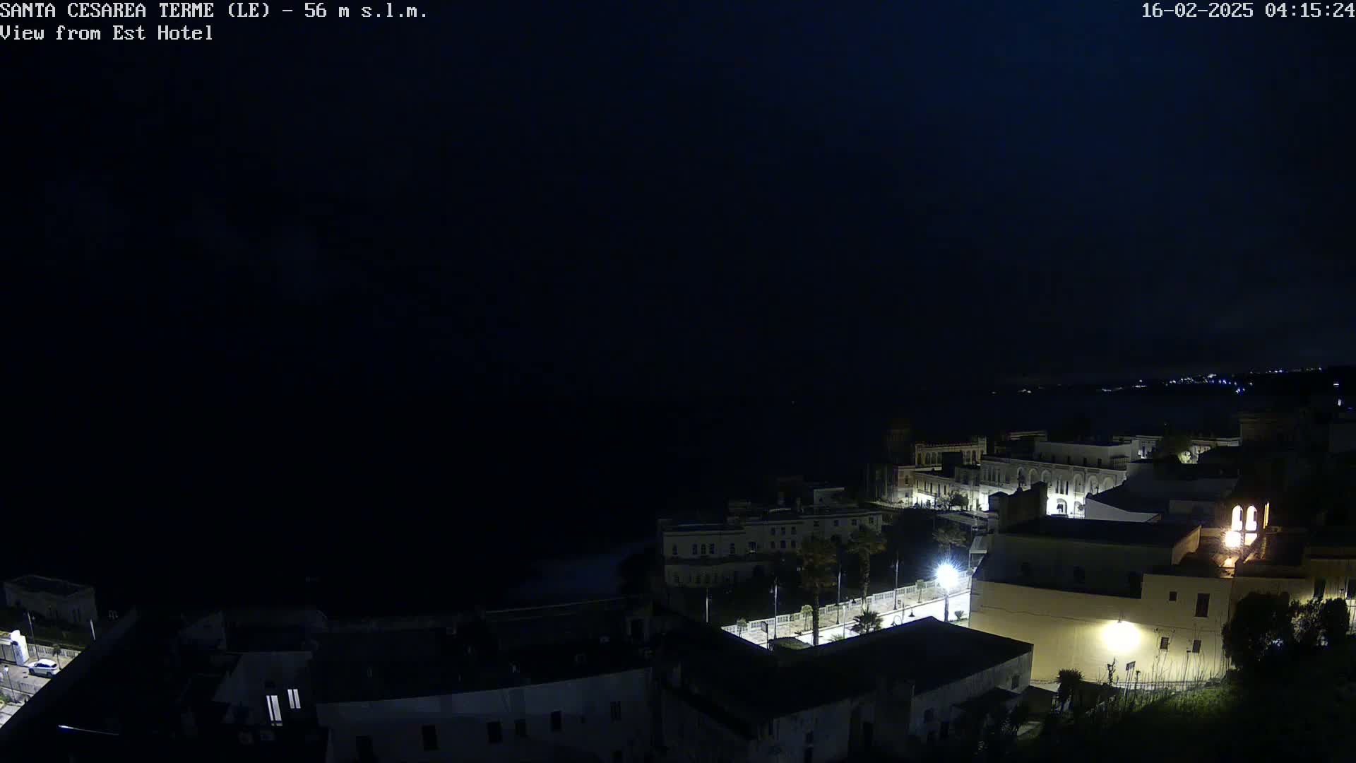 A nighttime, dark view of a coastal town with buildings illuminated by streetlights, overlooking a dark sea under a clear night sky.