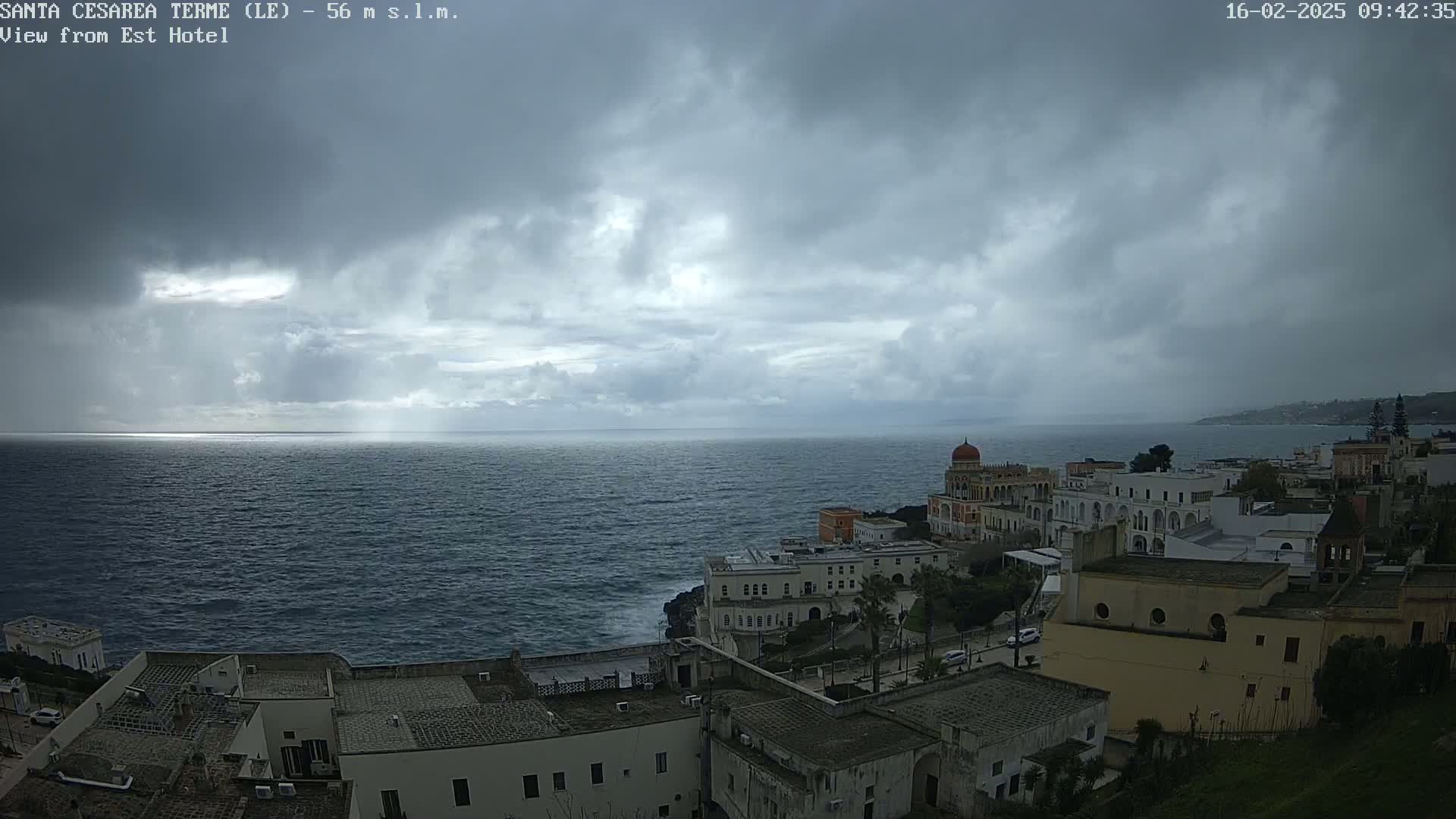 A coastal town with white buildings sits on a cliff overlooking a dark blue sea under a cloudy, overcast sky.