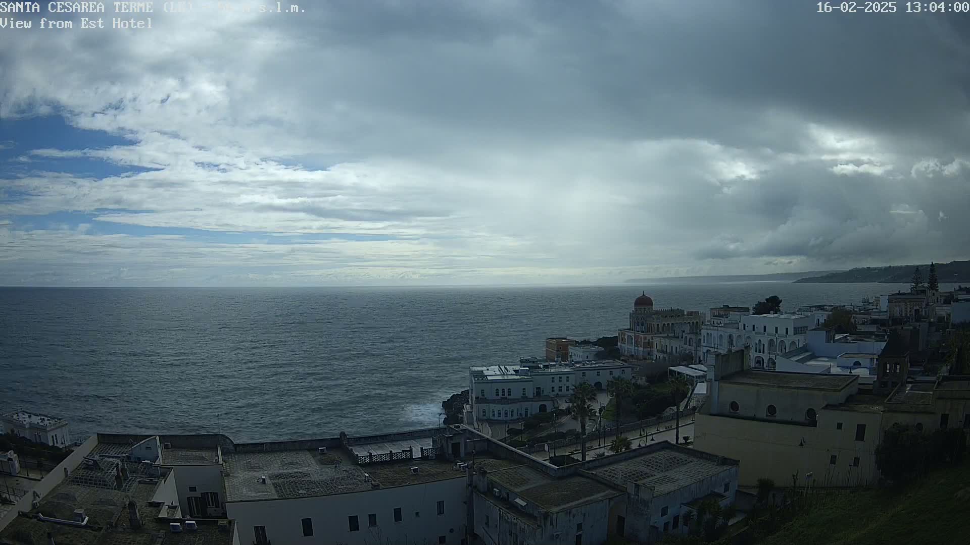 A coastal town with white buildings sits beneath a mostly cloudy sky, overlooking a dark blue sea.