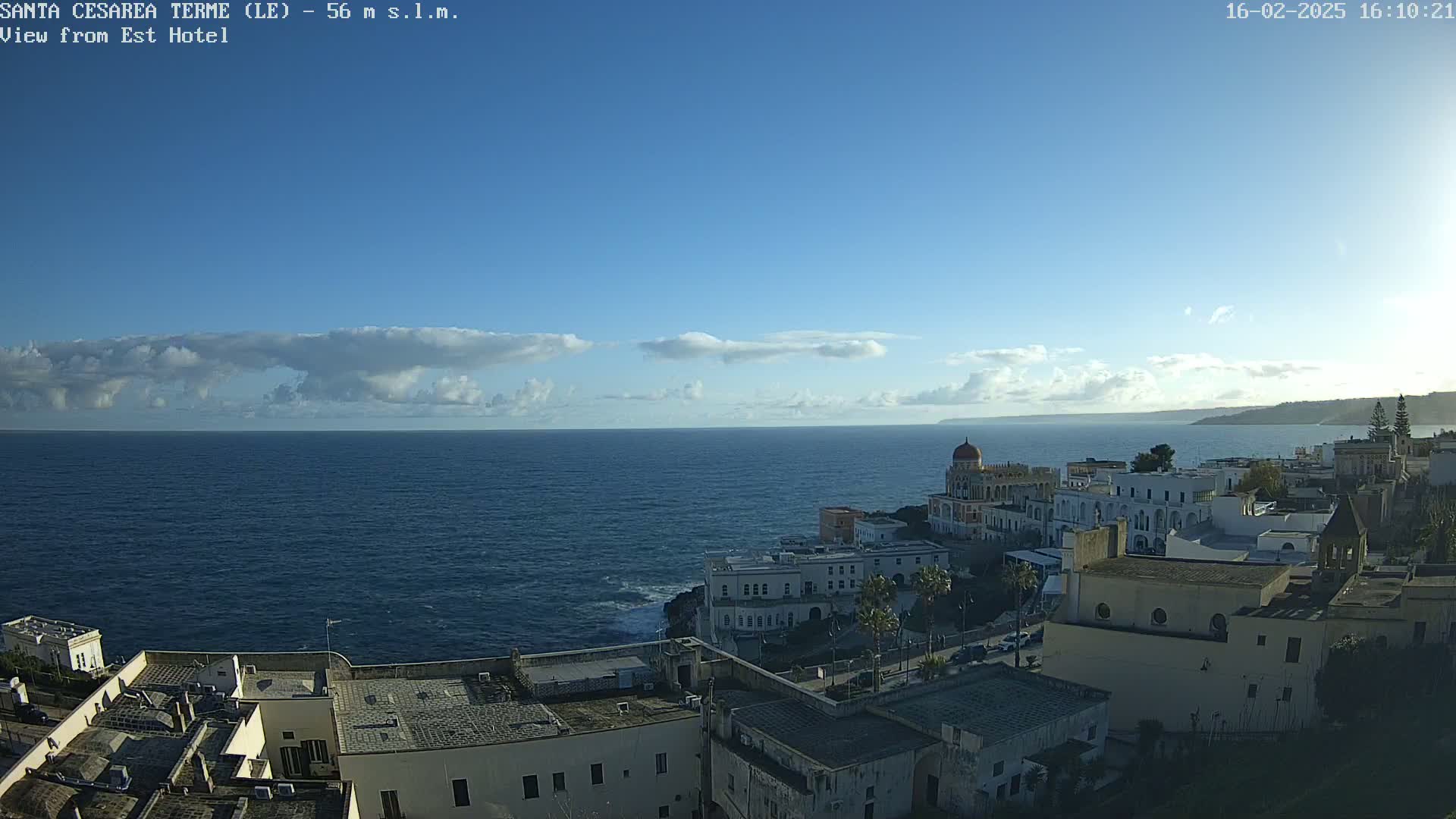 A coastal town with white buildings and a dome-shaped structure is situated on a rocky outcrop overlooking a calm blue sea under a mostly clear sky with some scattered clouds.