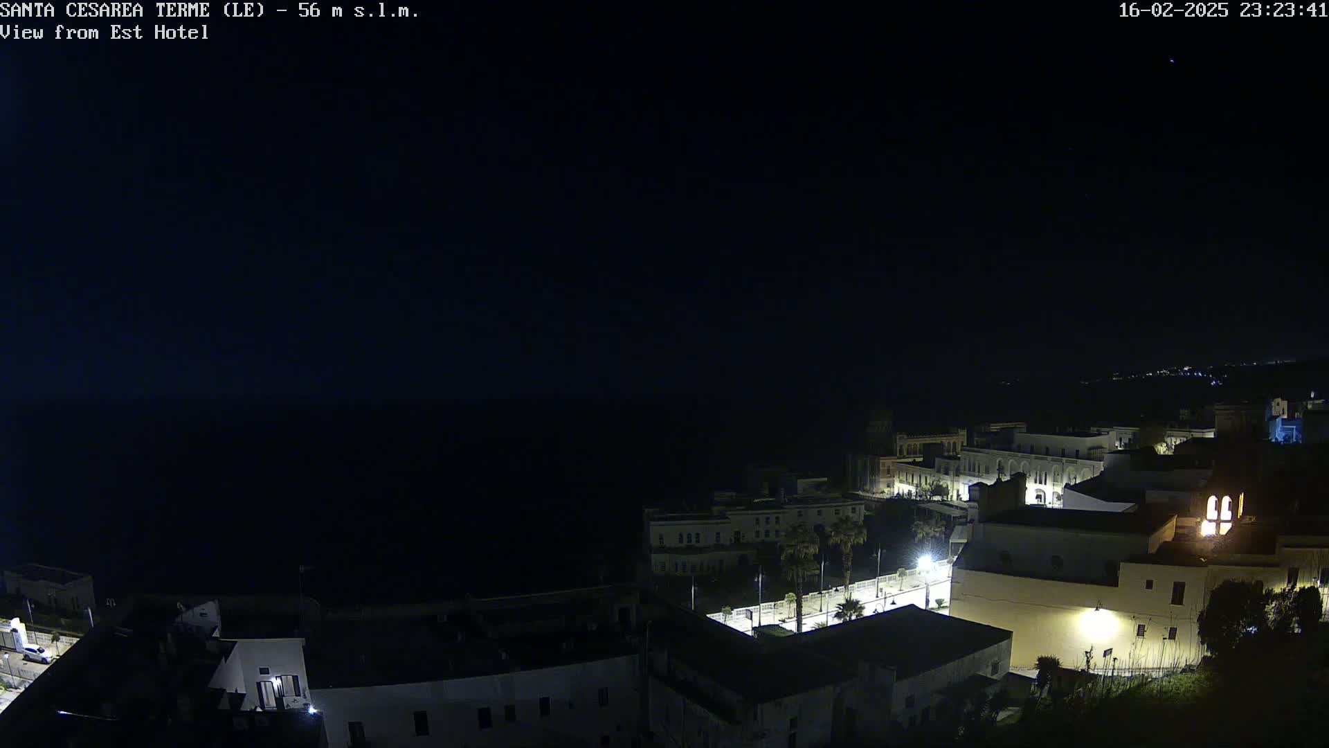 A nighttime, high-angle view of a coastal town, with dimly lit buildings near a dark body of water under a clear, starlit sky.