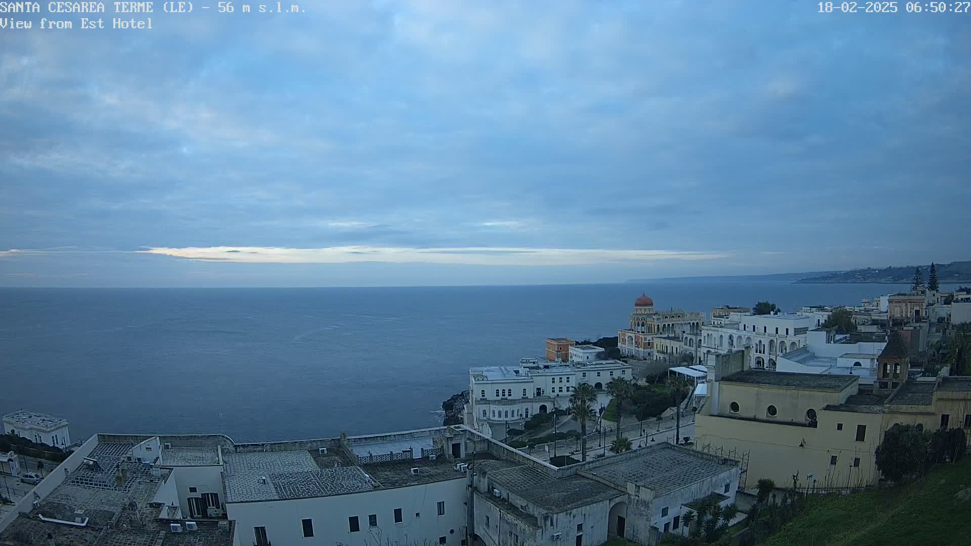 A coastal town with white buildings overlooks a calm, dark blue sea under a mostly cloudy, pale blue sky.