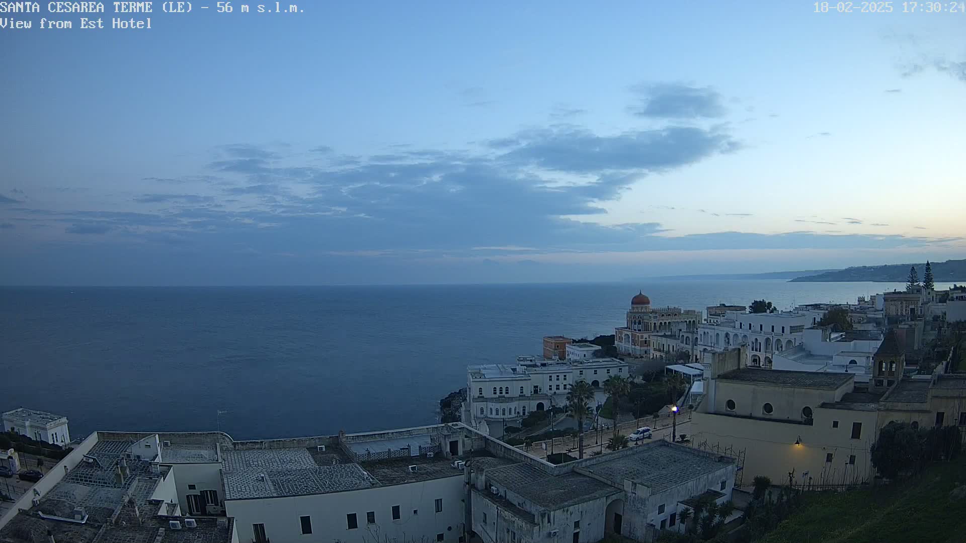 A coastal town with white buildings and a domed structure overlooks a calm, dark blue sea under a twilight sky with thin clouds.