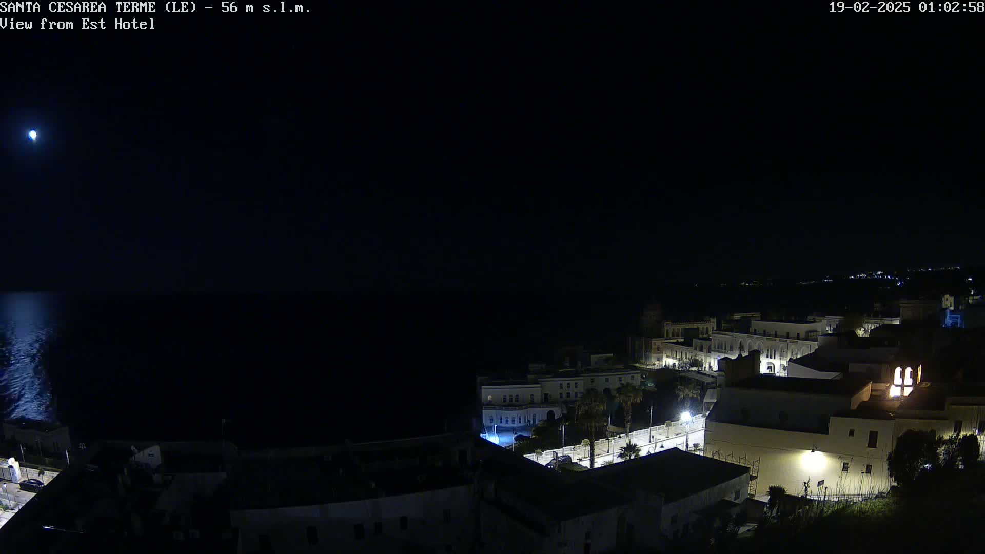 A nighttime, seaside town is partially illuminated, with a bright moon visible in a dark sky over a calm sea.