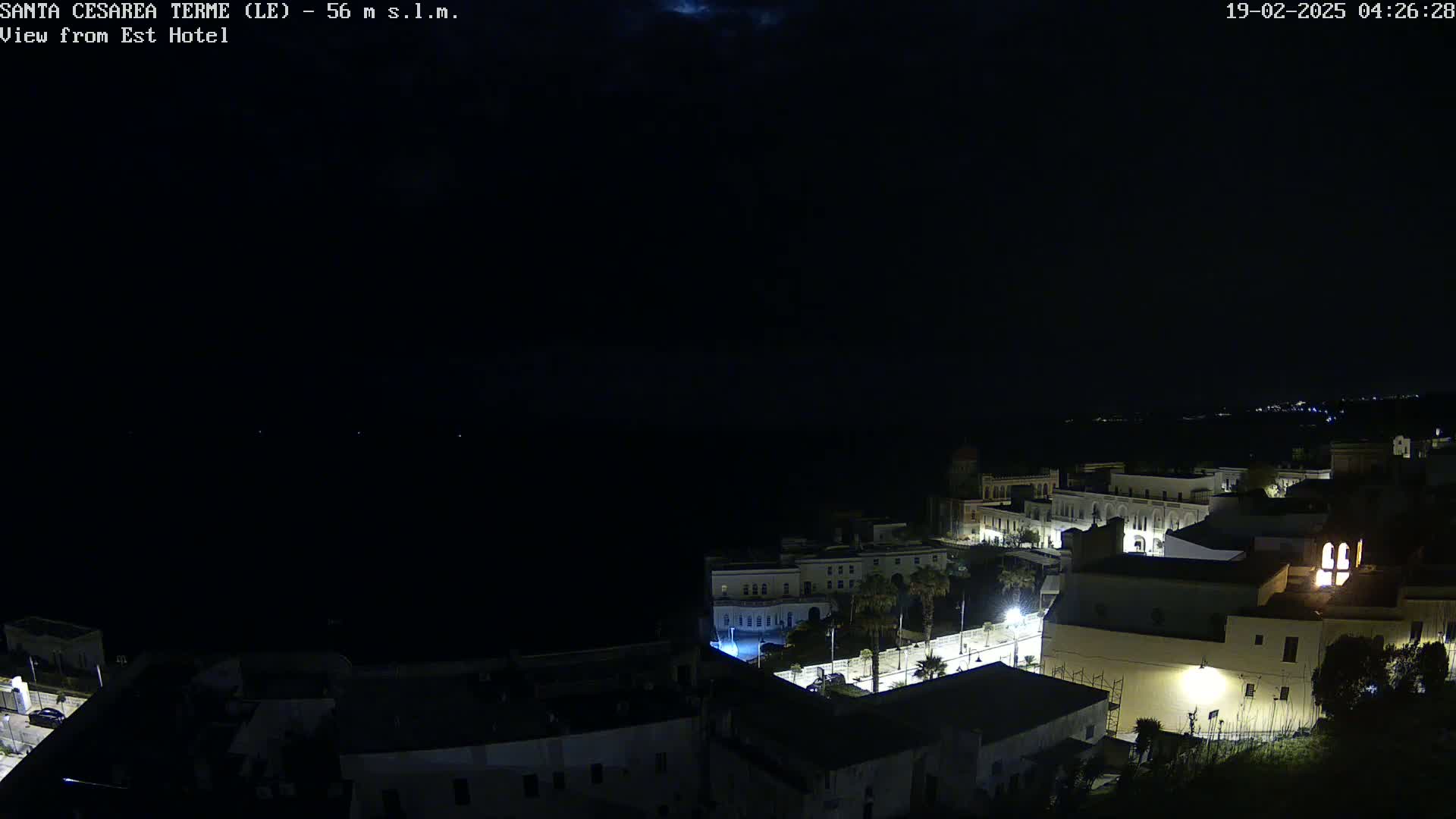 A nighttime, high-angle view of a coastal town with dimly lit buildings and a dark sea under a mostly clear sky.