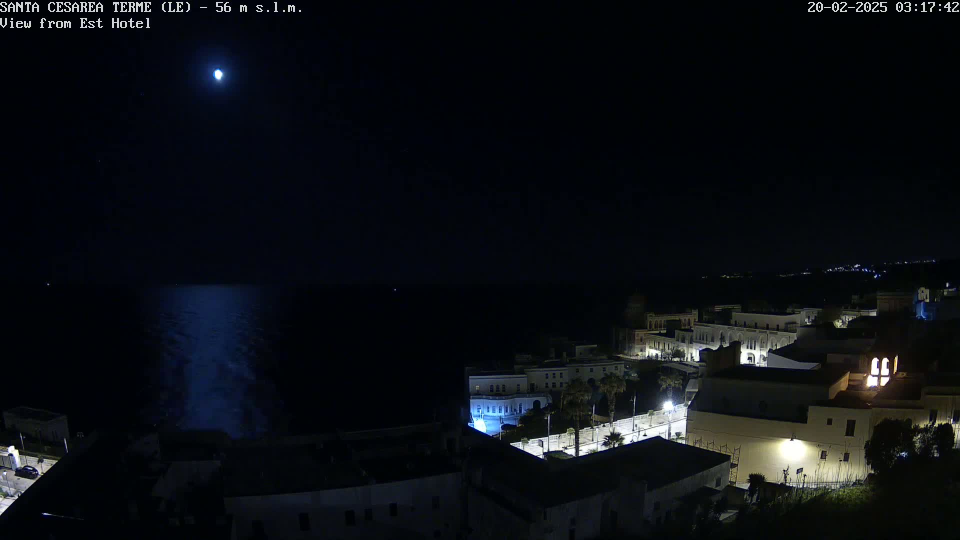 A nighttime view of a coastal town with a bright moon in a clear sky, overlooking a dark sea.