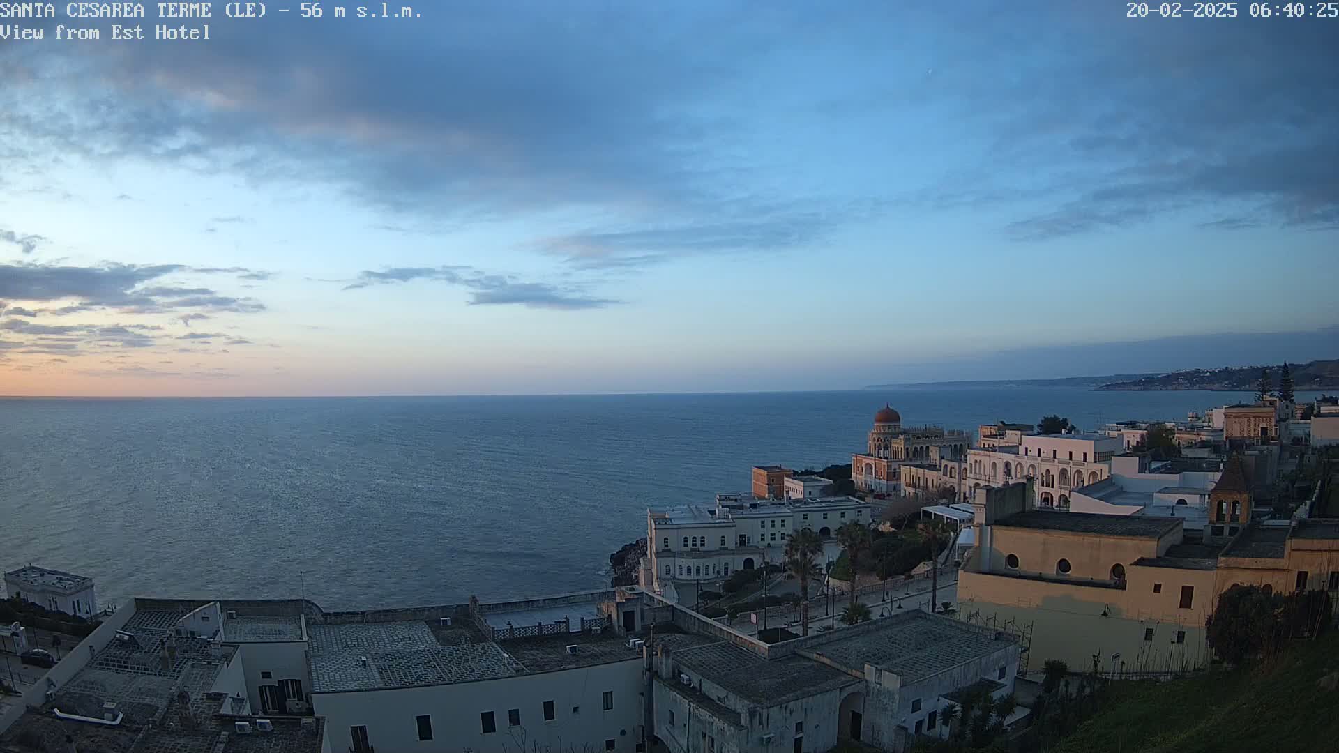 A coastal town with white buildings, a domed structure, and green vegetation overlooks a calm, dark-blue sea under a twilight sky.