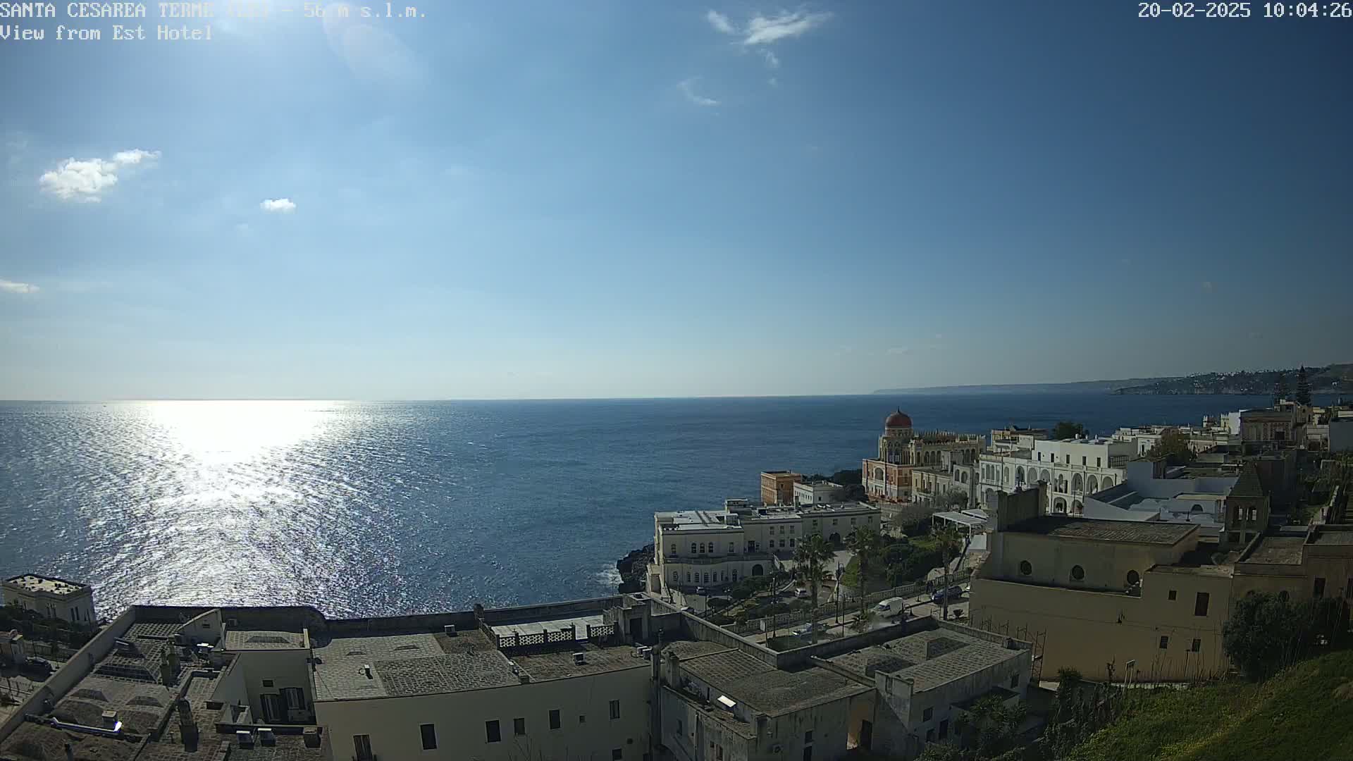 A coastal town with white buildings sits on a cliff overlooking a calm, sunlit sea under a mostly clear sky.